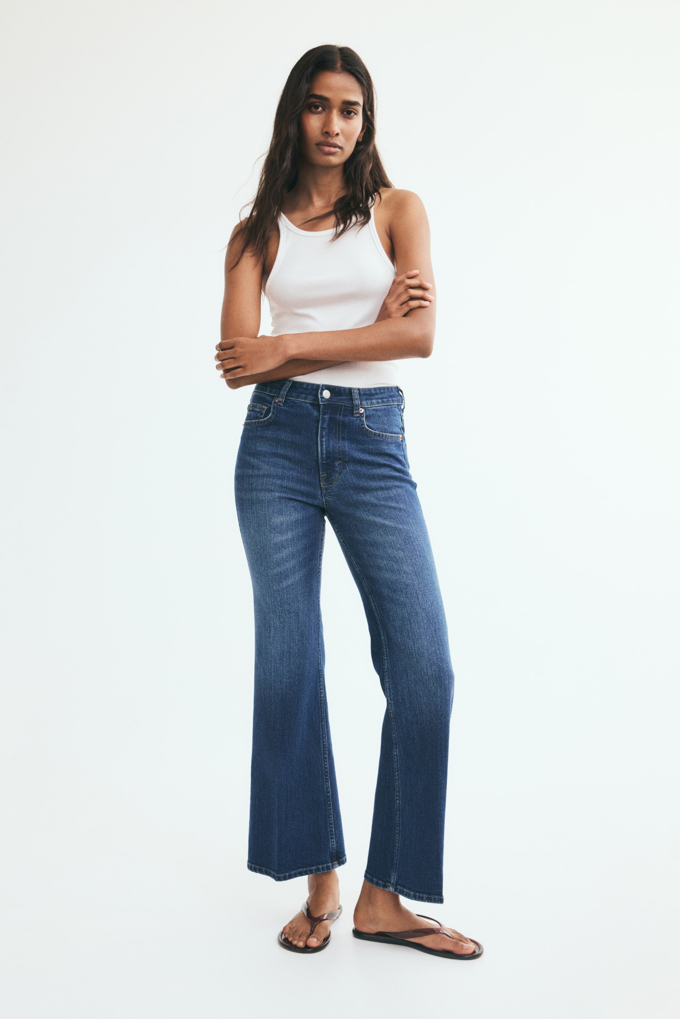 View larger image: Blue high-waisted jeans with a slim fit through the thighs, a flared cut from the knees, and subtle front creases are being worn by the model. The jeans are styled with a tucked-in white tank top and dark flat sandals.