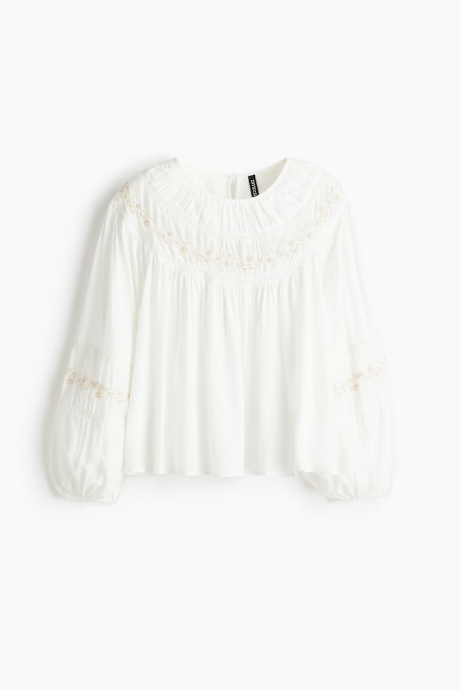 A white blouse in a viscose weave is being presented from the FRONT, featuring a round neckline with a keyhole opening and a button at the back. It is showcasing dropped shoulders and long balloon sleeves with buttons at the cuffs. The blouse is detailed with light-colored floral embroidery and lace insets around the neckline and on the sleeves. - 1