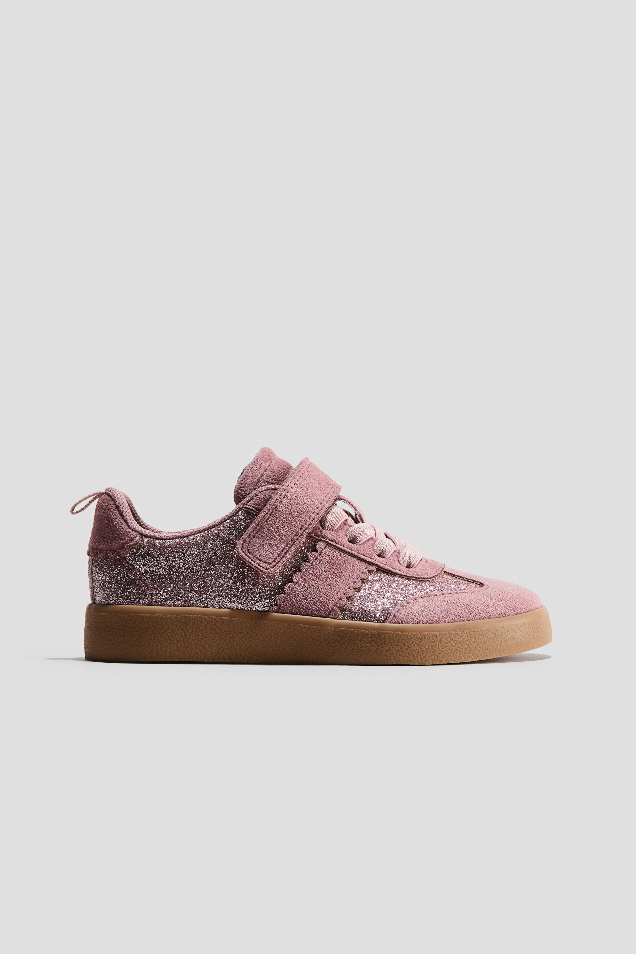 Xem ảnh lớn hơn: Một đôi sneaker màu hồng đang hướng sang phải, với các tấm bên lấp lánh, phần mũi giày, gót giày và khu vực xỏ dây có chất liệu giống da lộn, cùng viền lượn sóng răng cưa phân tách các bề mặt chất liệu. Một quai dán hook-and-loop đơn đang cố định phần thân giày bên trên dây buộc, và giày được đặt trên đế phẳng màu nâu.