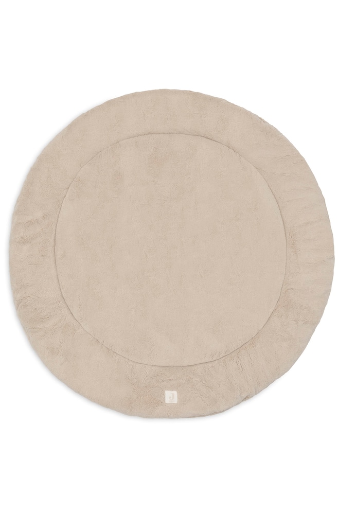 Un tapis de parc pour bébé rond, de couleur sable et moelleux, est posé à plat, mettant en valeur sa surface douce et texturée. Le tapis présente une bordure circulaire cousue ainsi quâune petite étiquette logo carrée sur le bord inférieur. - 1