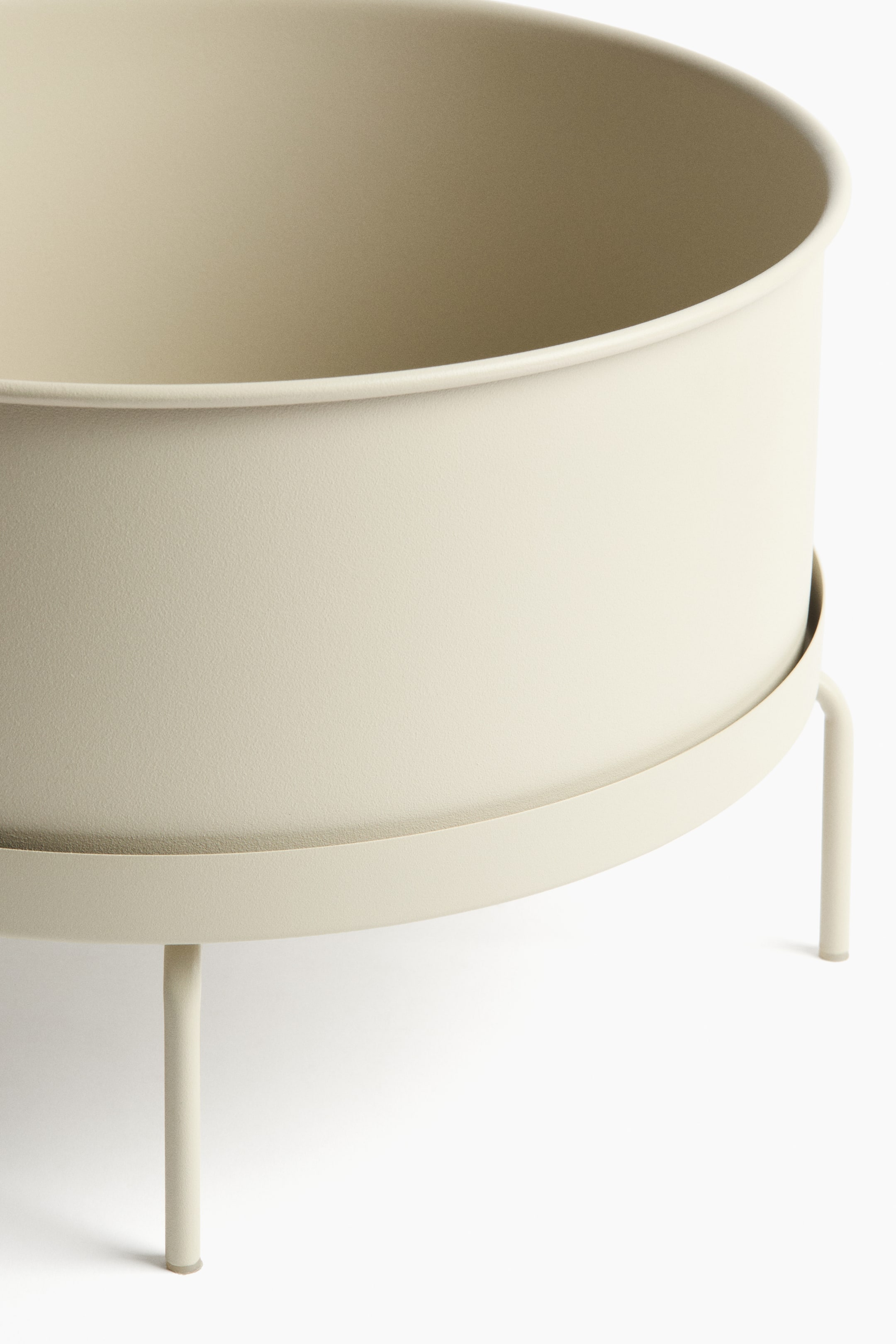 Ampliar la imagen: Una maceta de metal ancha y de color beige claro descansa sobre un soporte a juego, con el costado de la maceta visible por encima del platillo circular del soporte. El soporte presenta patas delgadas y cilíndricas.