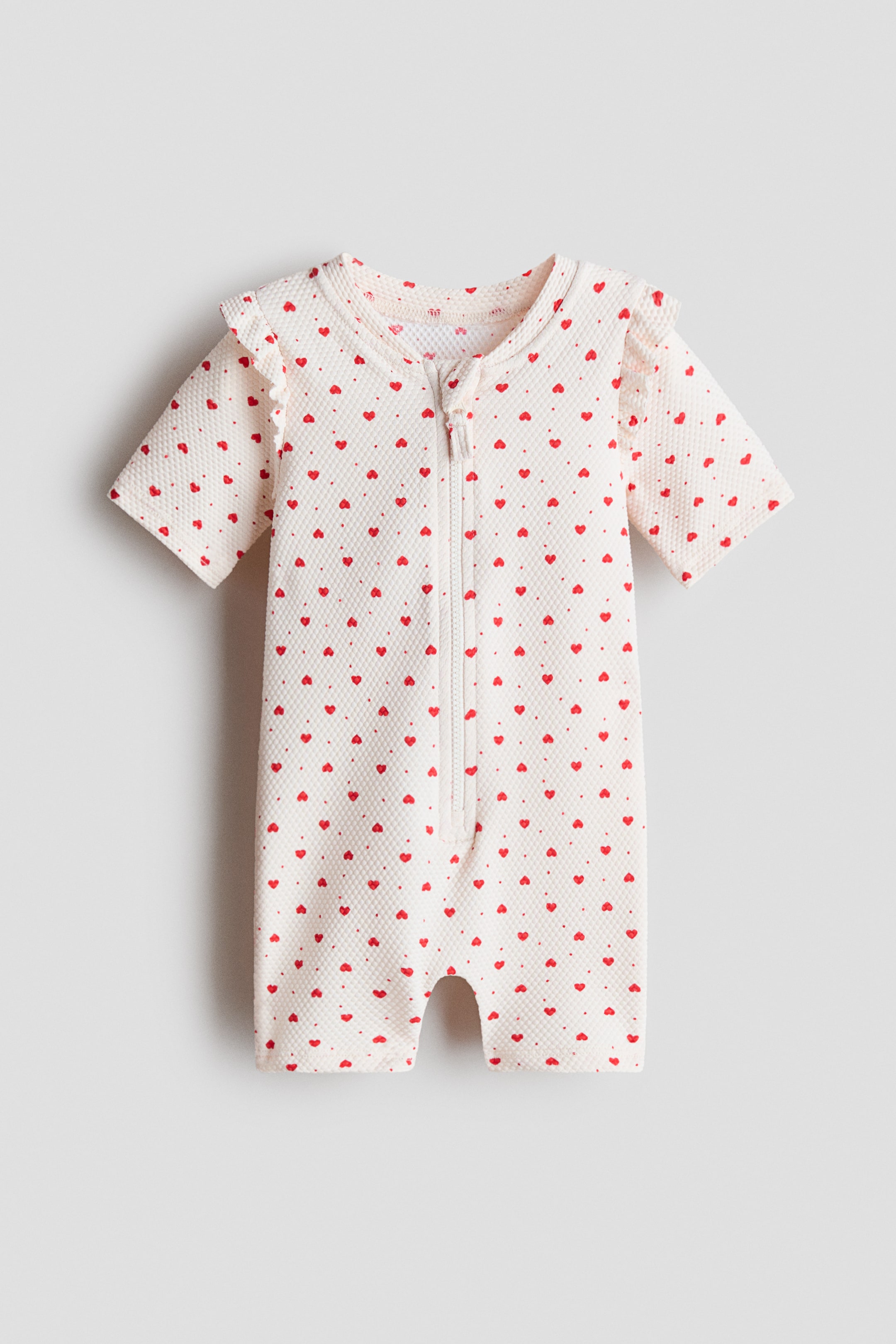 拡大画像を表示: クリーム色でテクスチャーのある水着が正面から見えます。小さな赤いハートとドットがパターンされており、ラウンドネックライン、肩にフリルのついた短い袖、短い脚、中央前部に沿って白いジッパーが付いています。