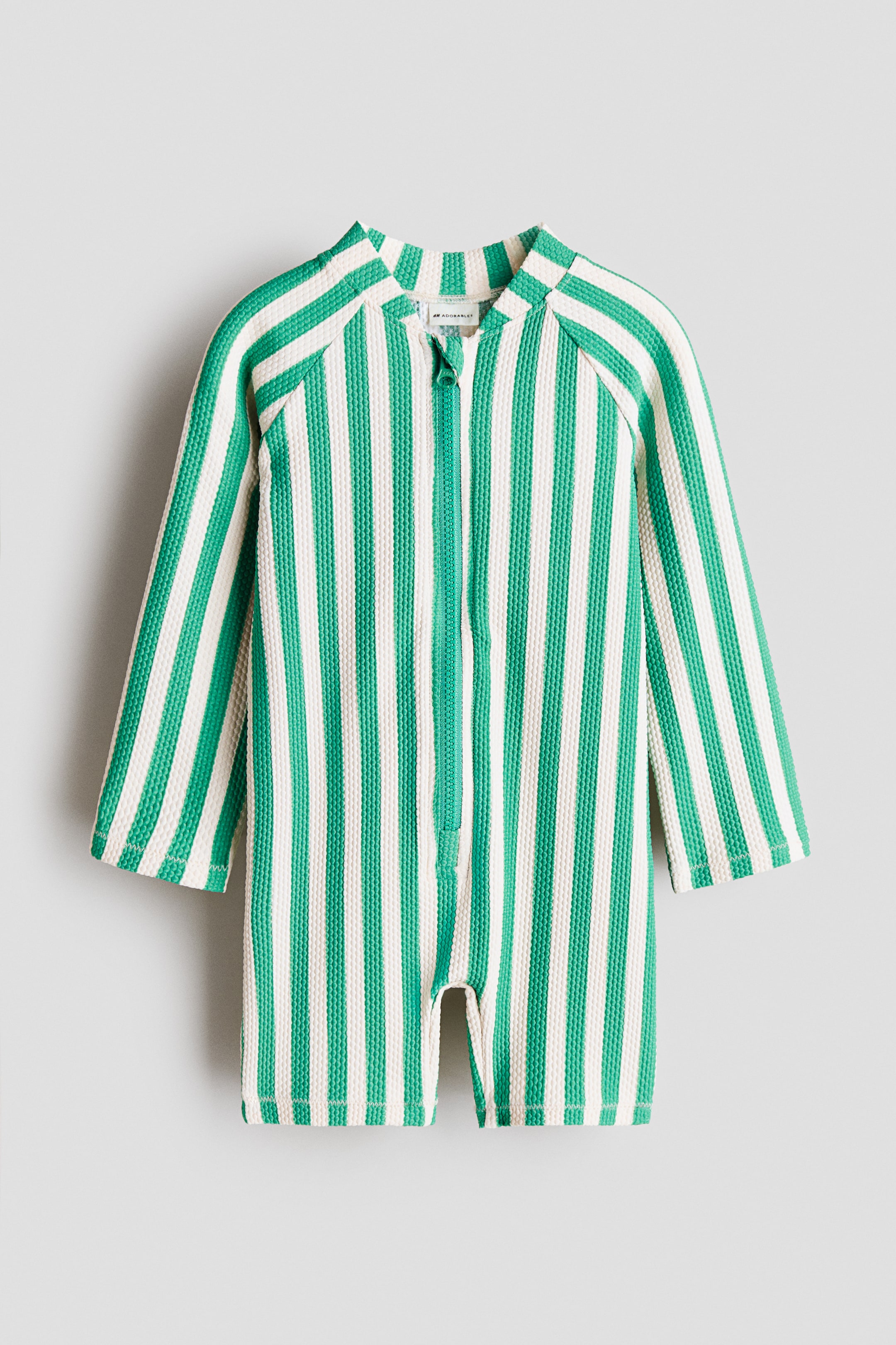 Voir une image plus grande: Un maillot de bain présenté de face, arborant un motif intégral de rayures verticales vertes et crème sur une matière texturée, un petit col montant, une fermeture éclair à l’avant, des manches raglan longues et des jambes courtes.