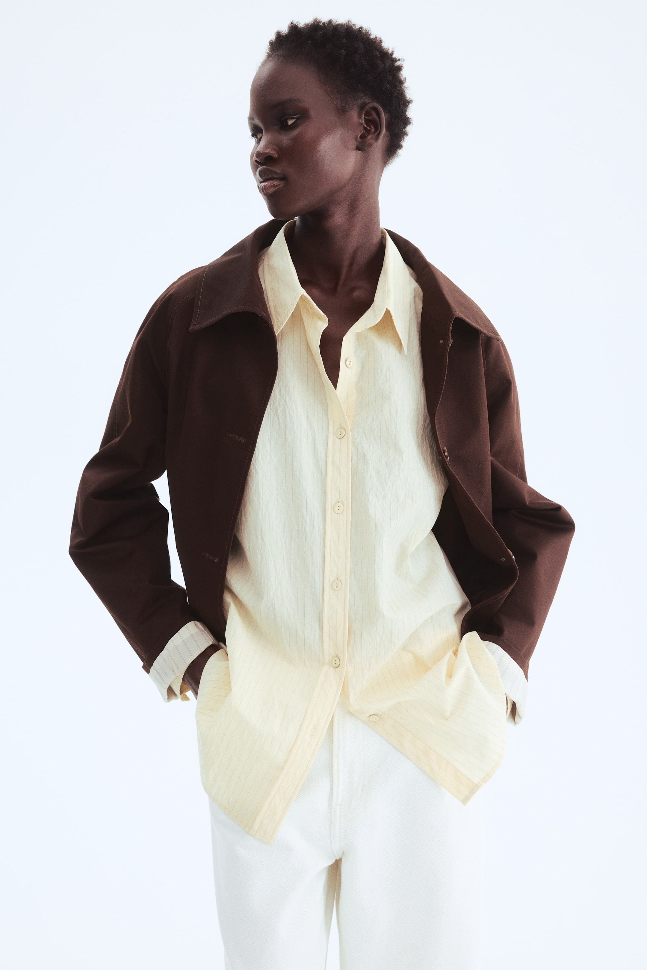 이미지 확대 보기: 어두운 갈색 재킷이 열려 있고, 그 아래로 밝은 노란색 버튼다운 셔츠가 드러나고 있습니다. 셔츠는 미세한 세로 줄무늬가 특징이며, 커프스가 재킷 소매를 넘어 연장되어 있습니다. 이 스타일은 흰색 바지와 함께 연출되었습니다.