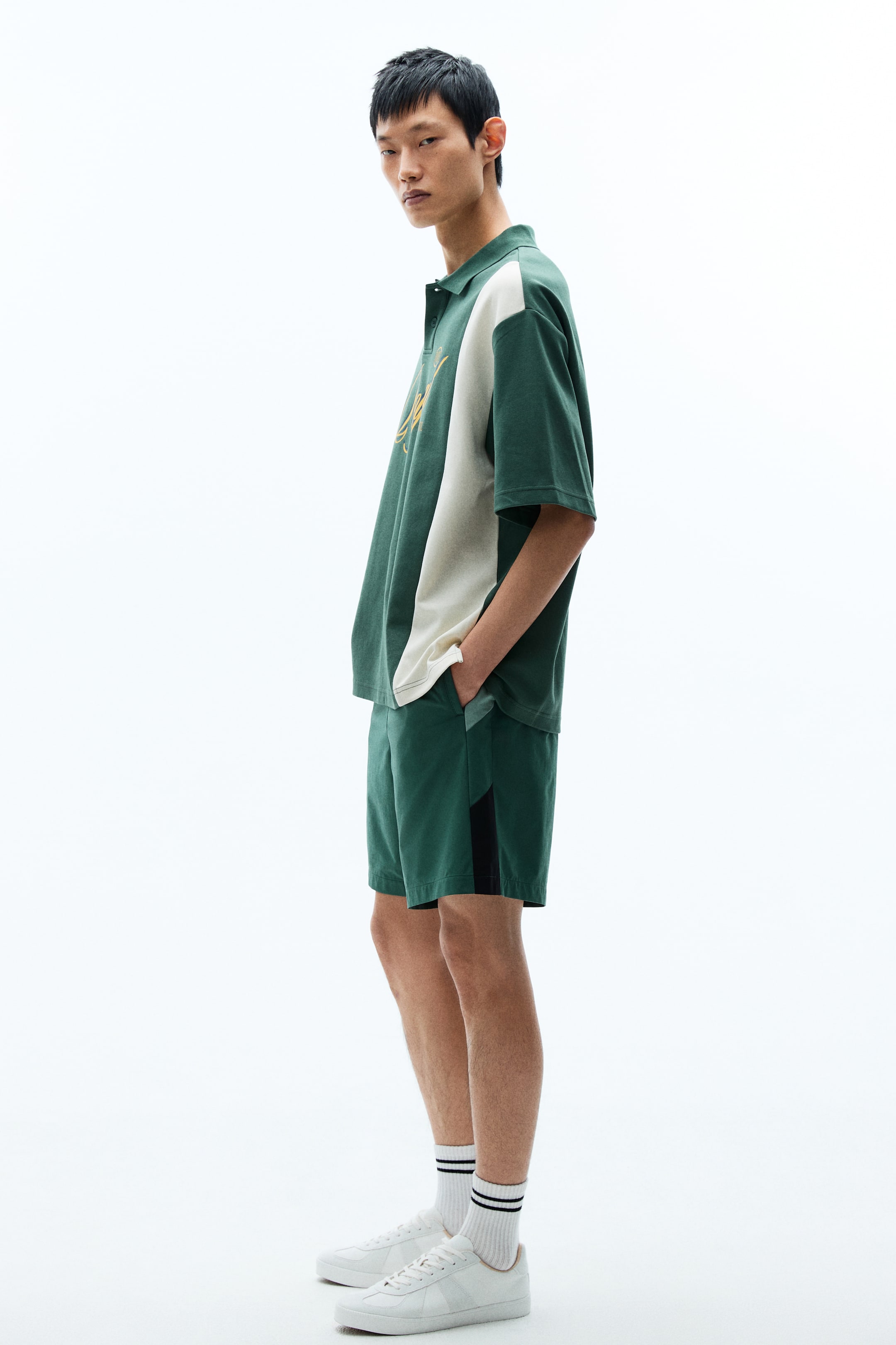 View larger image: A matching dark-green polo shirt and shorts set is shown in side profile. The polo features a cream-coloured vertical panel on its right side and gold-embroidered script lettering on the chest, while the coordinating dark-green shorts display a black vertical stripe along the outer side of the left leg.