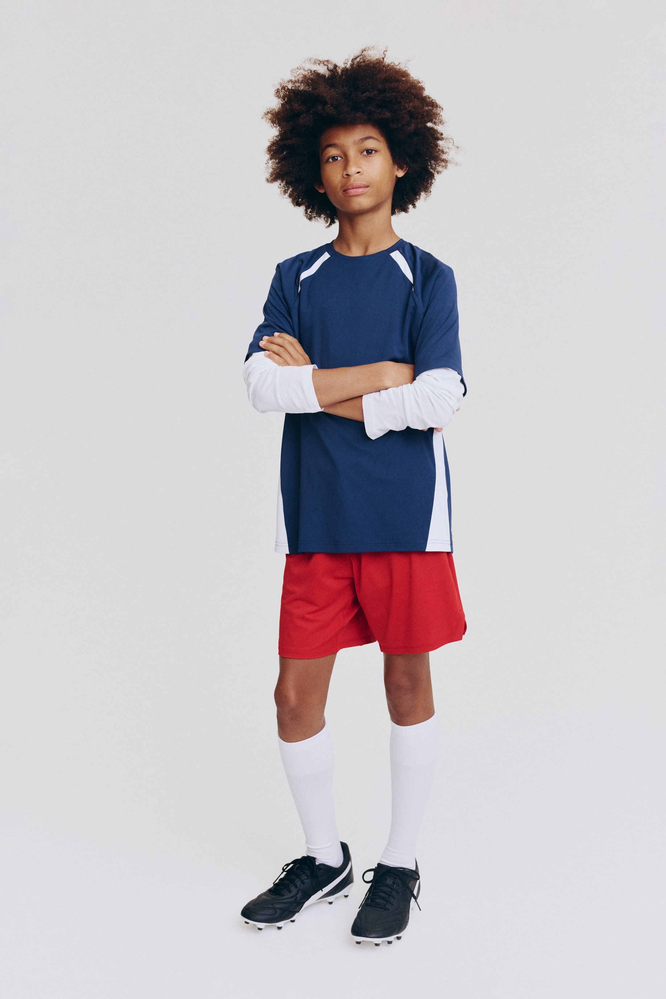 Größeres Bild ansehen: Die Sportbekleidung ist von vorne zu sehen und besteht aus einem dunkelblauen Kurzarmtrikot mit weißen Raglan-Schultereinsätzen und weißen Seiteneinsätzen, darunter erscheinen weiße lange Ärmel. Dazu passen rote Sportshorts, weiße knielange Socken und schwarze Fußballschuhe mit weißen Akzenten.
