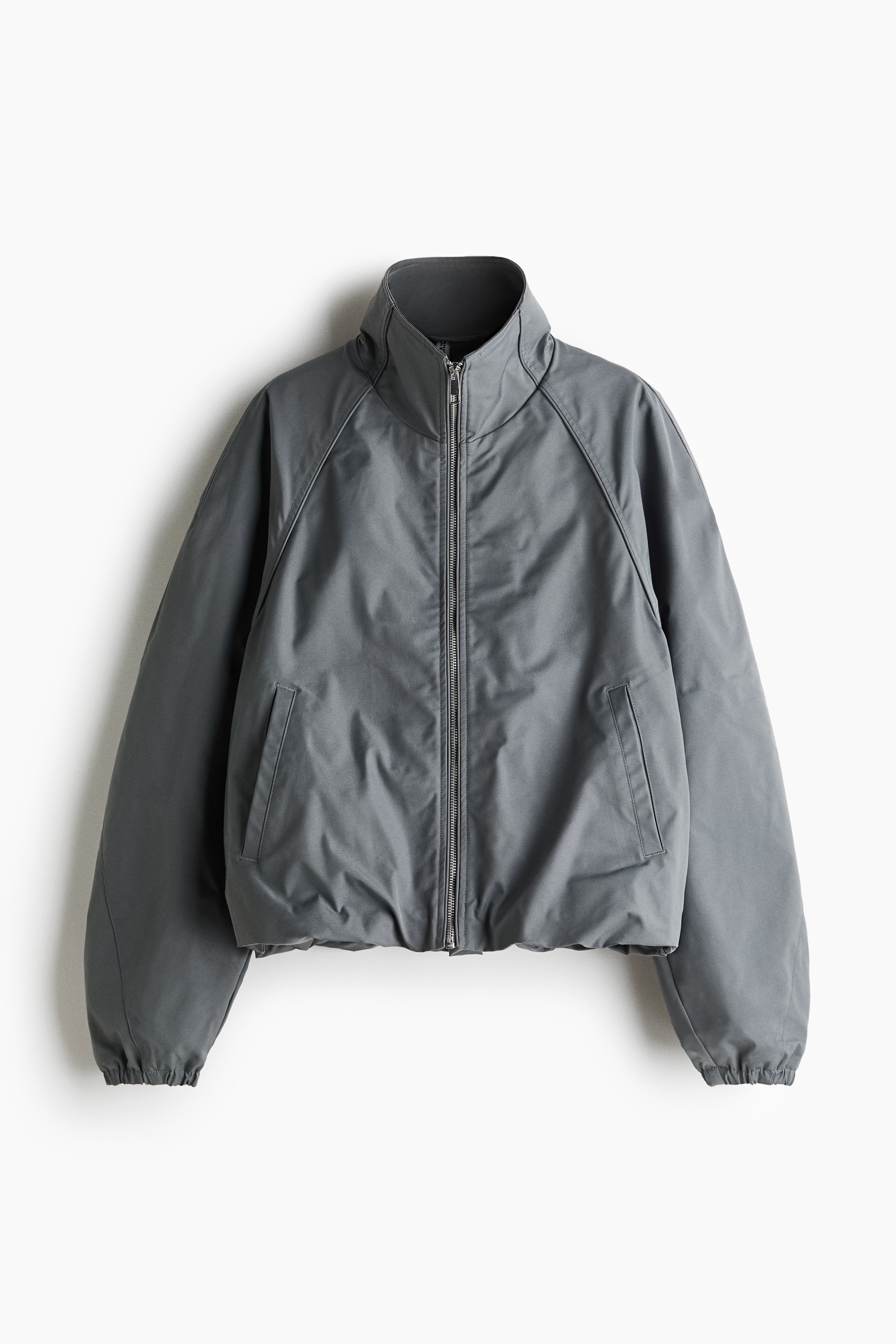 A dark grey oversized woven jacket is worn by the model, with their hands resting in the diagonal welt pockets. The jacket features a high stand collar, a front zip, and long raglan sleeves with elasticated cuffs.
