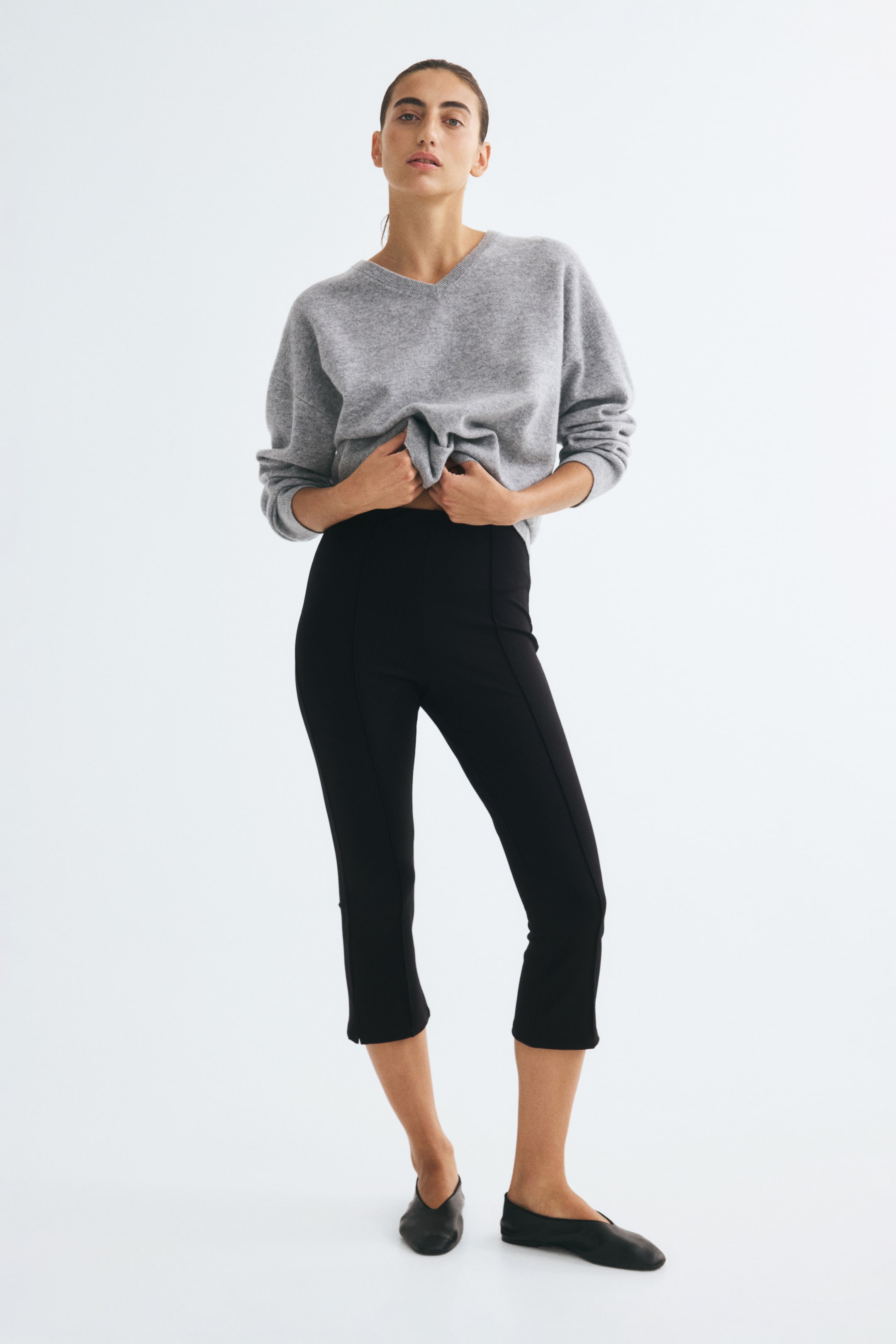View larger image: Black, 3/4-length fitted trousers with sewn-in front creases and small hem slits are worn by the model. The model is holding the hem of a grey knit sweater upward to reveal the high, elasticated waistband of the trousers, which are paired with black flat shoes.