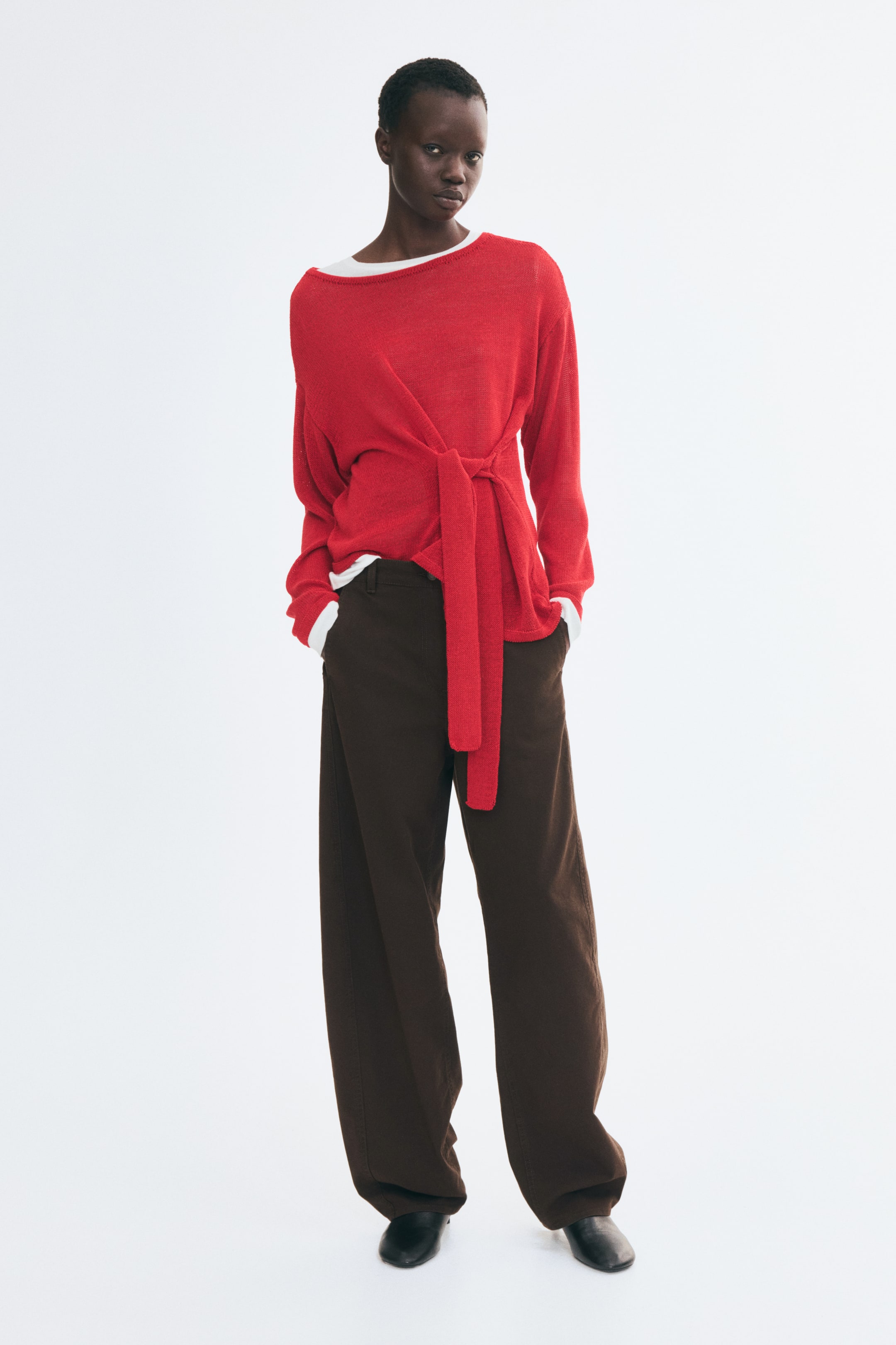 View larger image: The model is wearing dark brown, high-waisted barrel-leg pants and standing with her hands resting in the diagonal side pockets. The pants are styled with a red long-sleeve knit top featuring a waist-tie detail, a white undershirt, and black shoes.