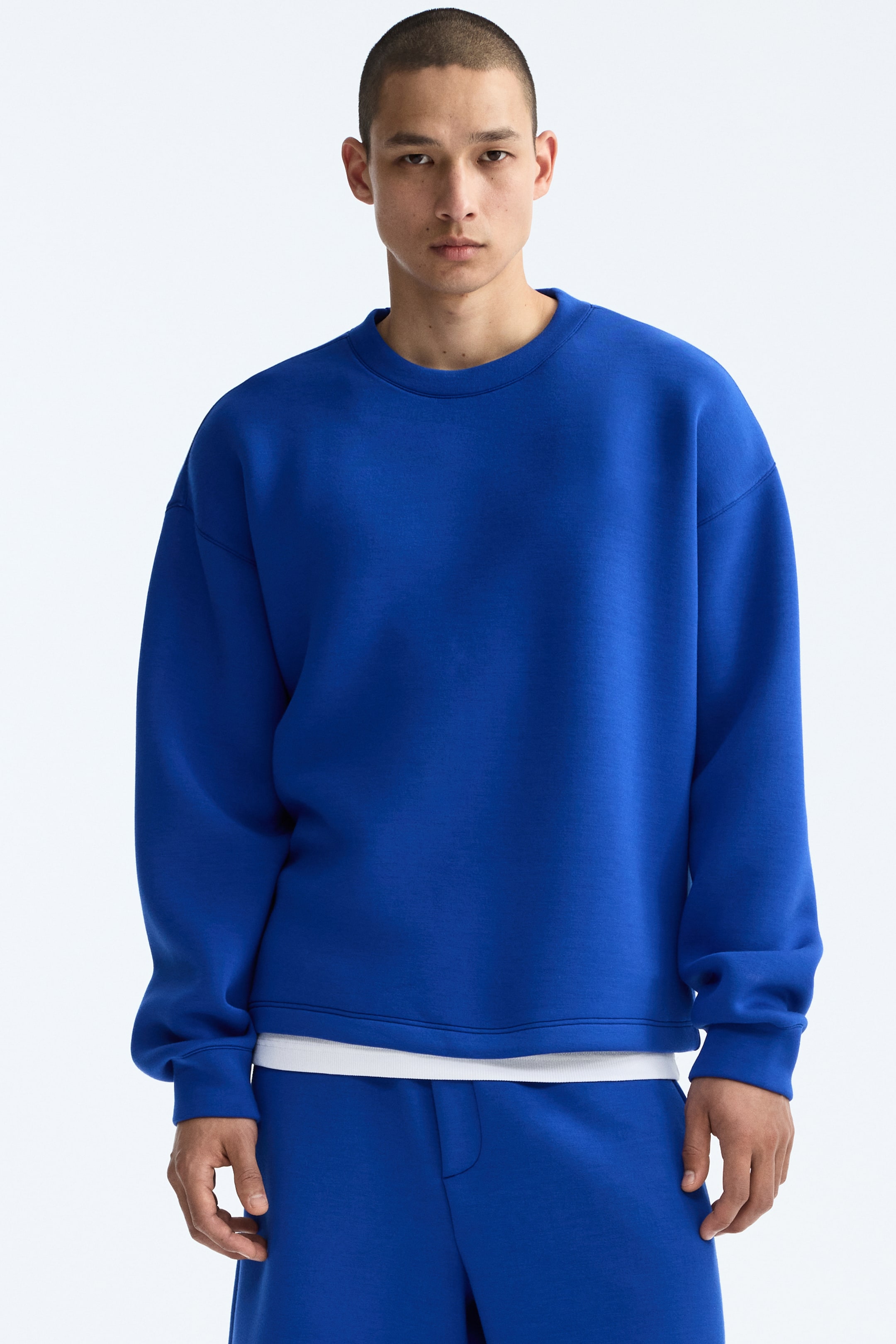View larger image: This blue sweatshirt showcases a crew neck, dropped shoulders, long sleeves with banded cuffs, and a banded hem. It's styled over a white t-shirt that visibly extends below the hem of the sweatshirt.