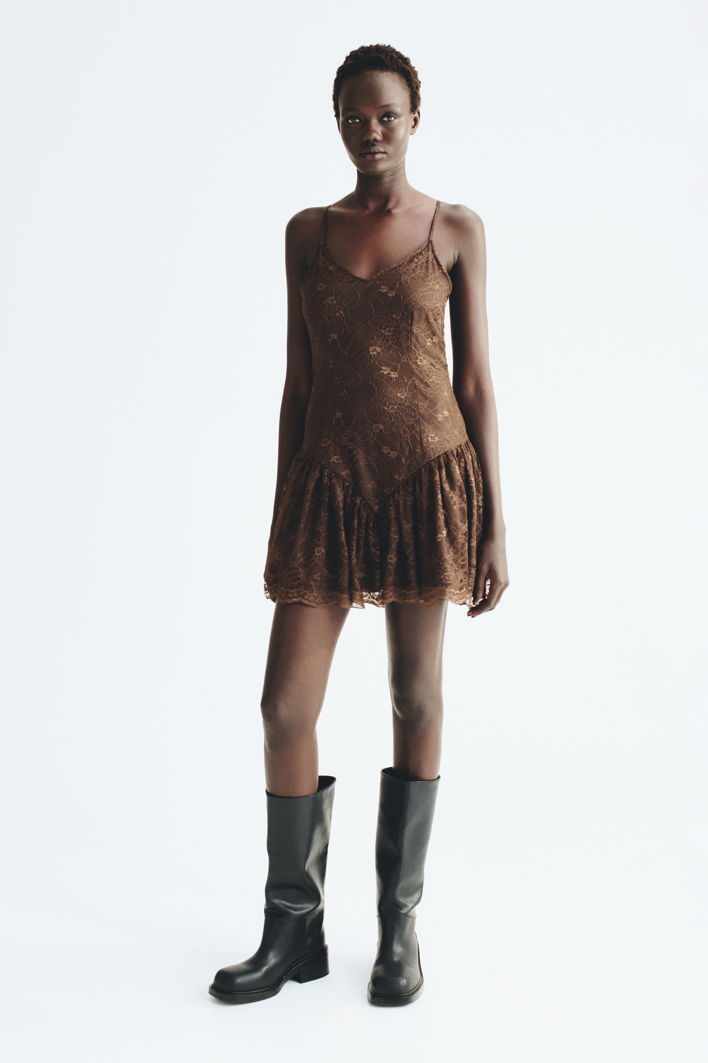 A brown lace mini dress is shown from the front, featuring adjustable spaghetti straps, a V-neckline, and a fitted bodice. The bodice extends into a dropped V-shaped waistline, which connects to a gathered, ruffled skirt with a scalloped hem.