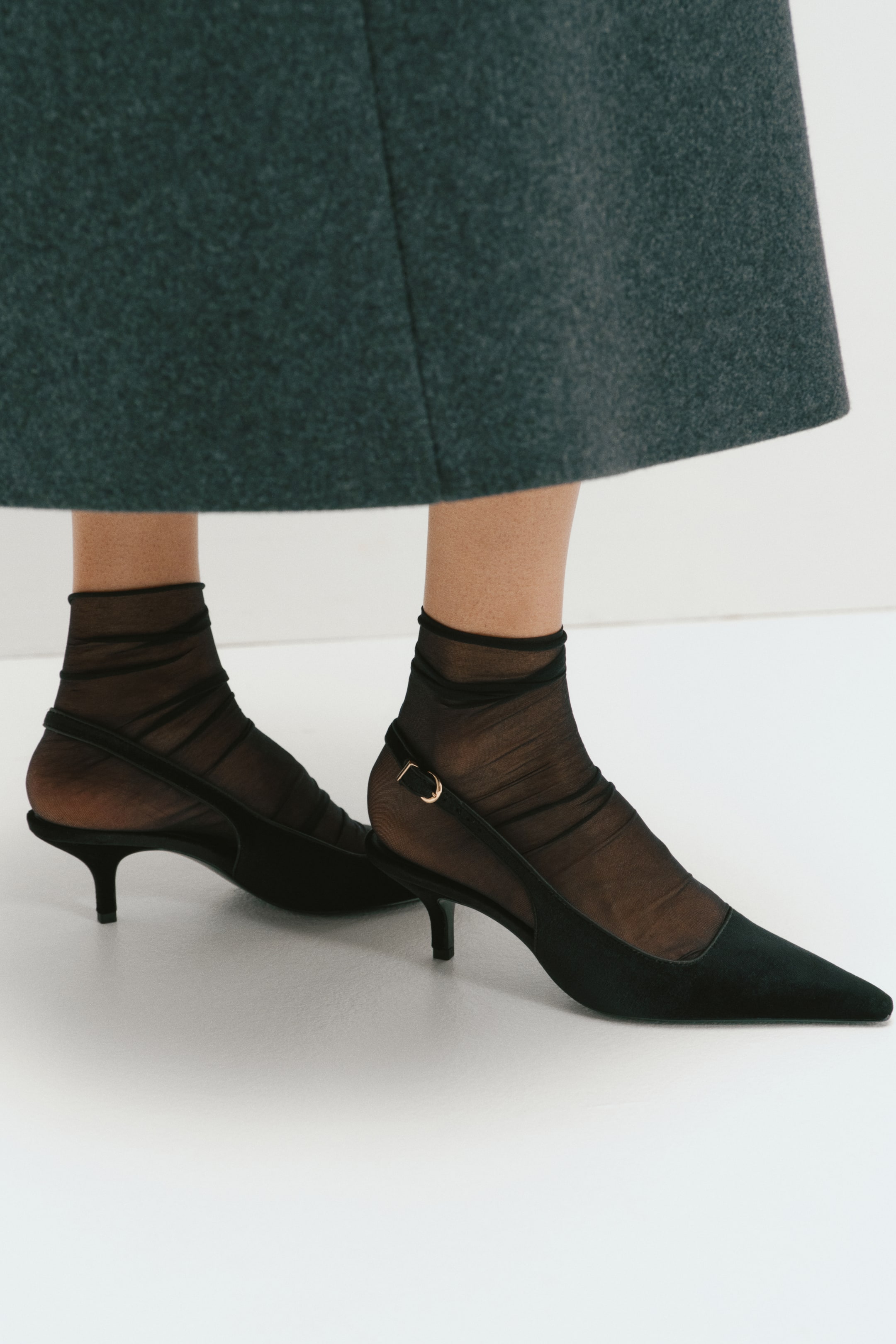 Größeres Bild ansehen: Schwarze Slingback-Schuhe mit spitzen Zehen, niedrigen Stilettoabsätzen und schmalen, verstellbaren Fersenriemen mit goldfarbenen Metallschnallen werden vom Model getragen. Die Schuhe werden mit transparenten schwarzen Knöchelsocken und einem dunkelgrauen Wollrock kombiniert.