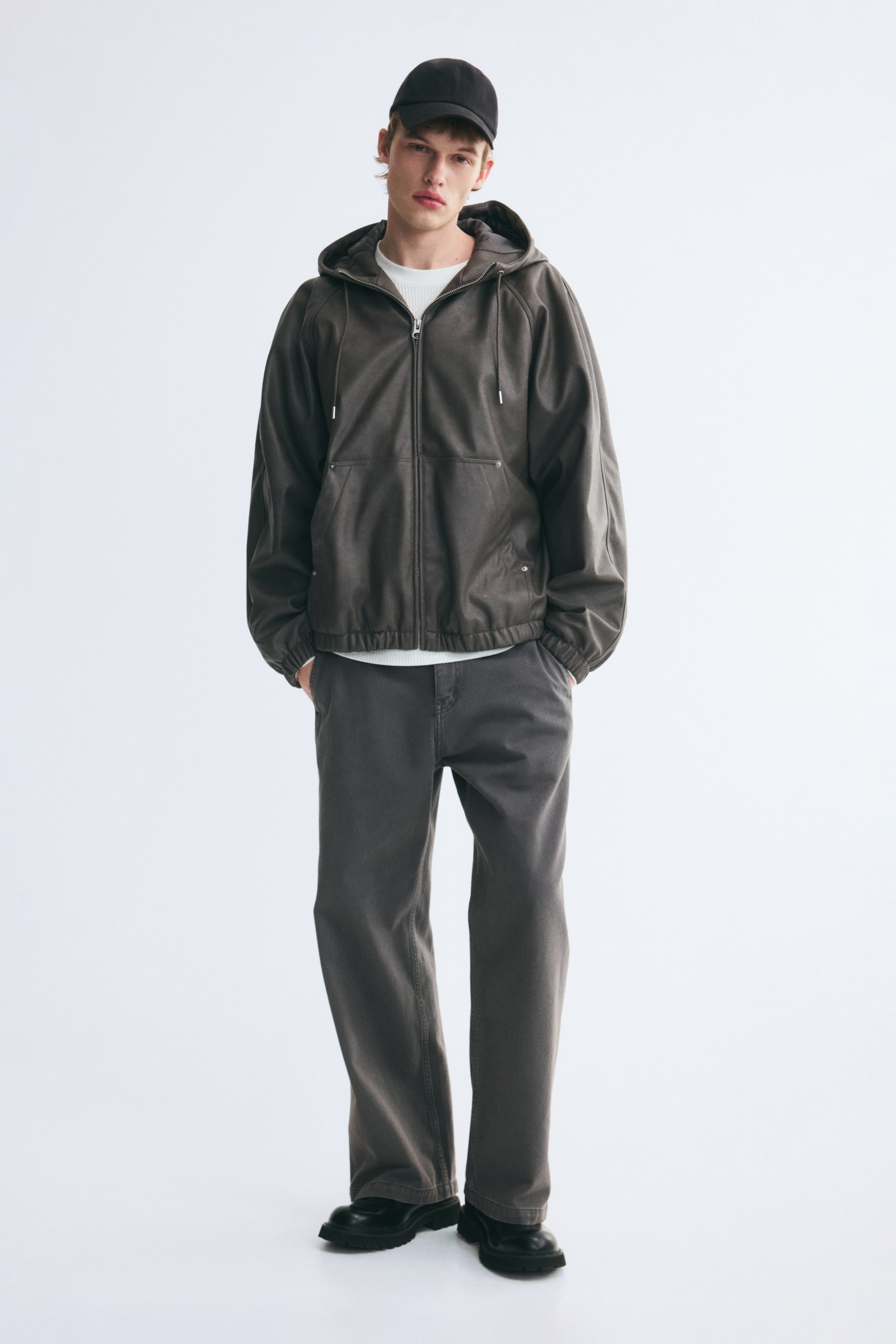 Größeres Bild anzeigen: Der/die Model trägt eine Carpenter-Hose aus dunkelgrauem Baumwoll-Twill mit lockerer Passform und weitem Bein sowie schrägen Seitentaschen. Das Model steht mit den Händen in den Seitentaschen und kombiniert die Hose mit einer dunkelbraunen Kapuzenjacke, einem weißen T-Shirt und schwarzen Stiefeln.