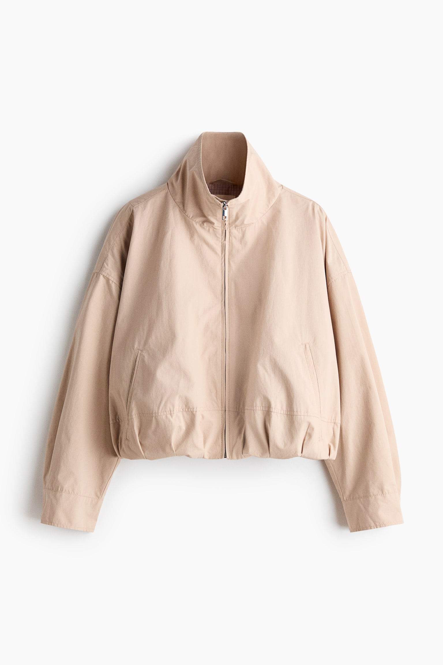 A beige jacket is presenting a front view, featuring a high stand-up collar and a silver-toned front zipper. It is showing wide, long sleeves with plain cuffs, an elasticated hem, and two front welt pockets. - 1
