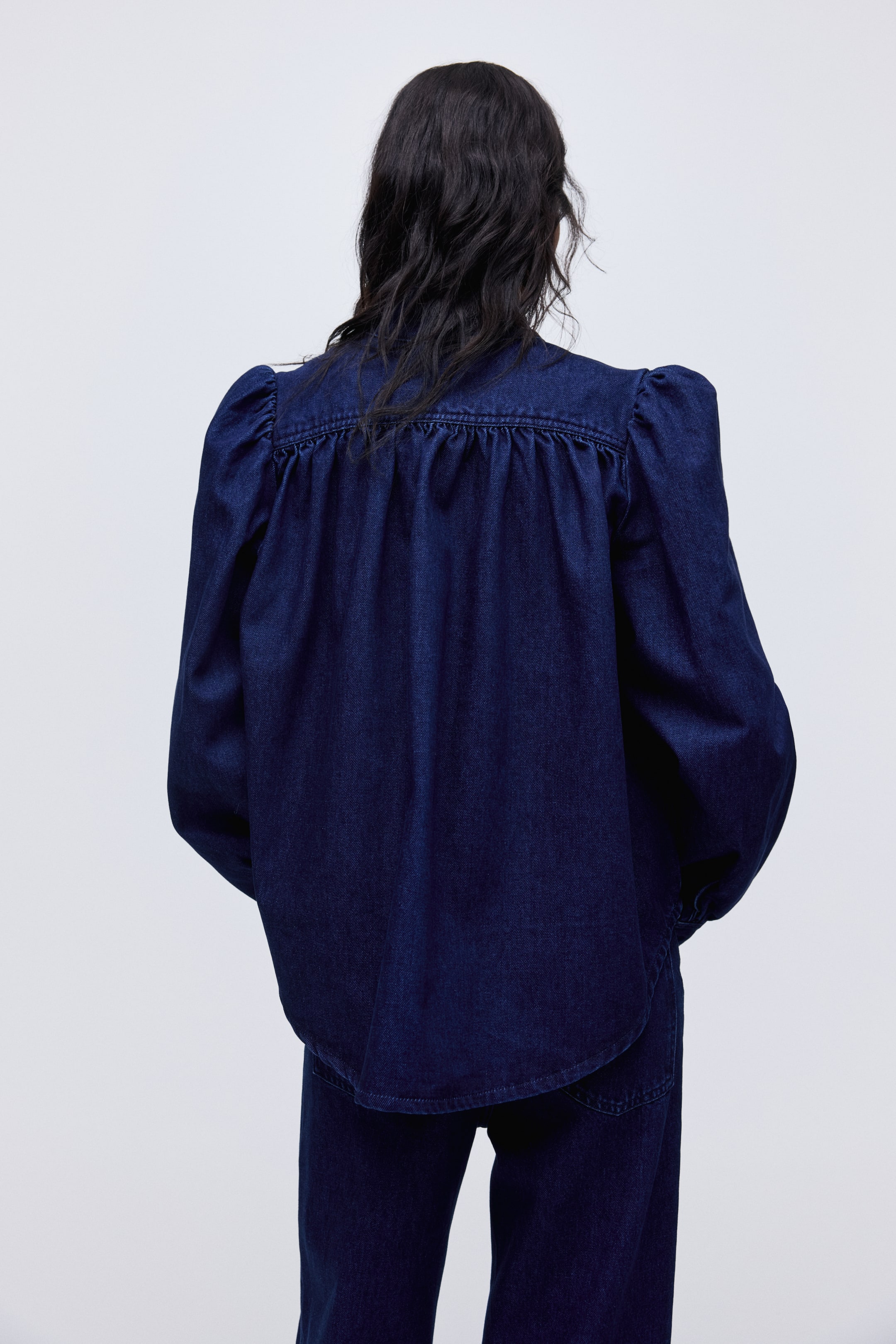 View larger image: The description remains the same, as it is already in English and adheres to US English standards: 

A dark blue denim top is presenting its back view, featuring a gathered back yoke with shirring below, creating fullness through the body, and long sleeves with gathered puff shoulders.