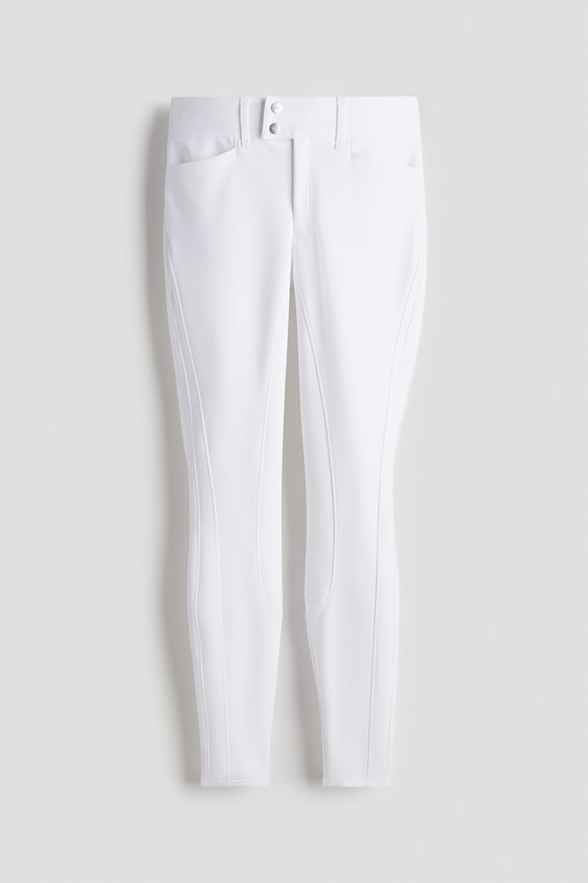 White breeches are being presented from the FRONT, featuring a high waist with an extended waistband, two decorative buttons, and visible front pockets. Textured silicone knee grips are accenting the inner knee areas, and elasticated binding is finishing the hems. - 1
