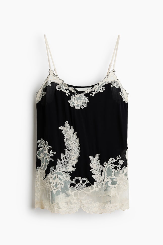 A black camisole is shown from the front, featuring adjustable spaghetti straps and a V-neckline trimmed with cream-coloured lace. Elaborate cream lace floral and leafy appliquÃ©s are climbing up the sides and accenting the sheer, scalloped hem. - 1