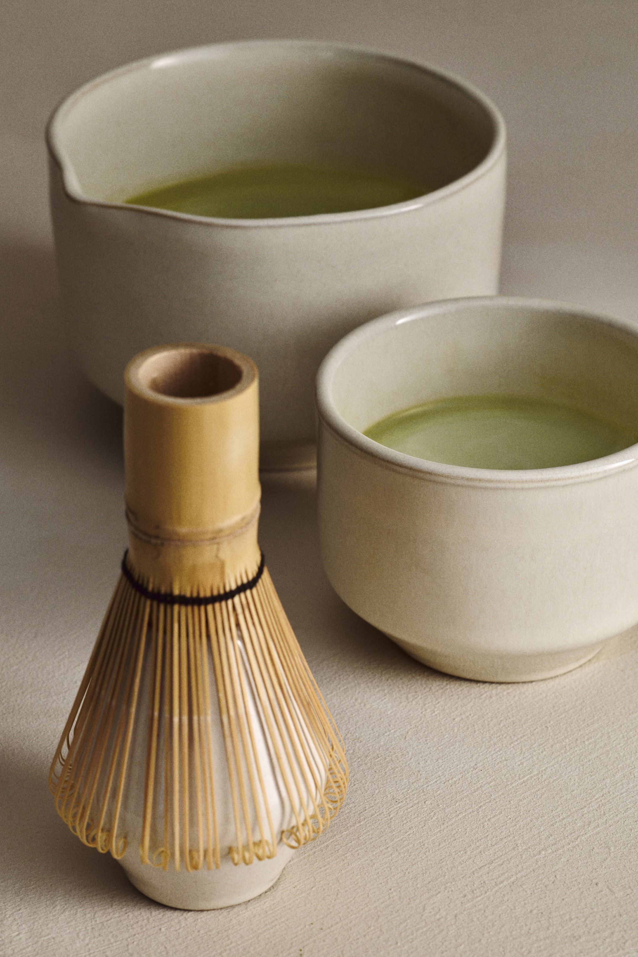 Näytä suurempi kuva: Bambuiset matcha-vispilä seisoo valkoisella keraamisella chasen-telineellä, hieman katsojaa kohti kallistettuna ja esittelee hienoja piikkejään sekä tummaa sidontaa. Suurempi vaalea keraaminen kaatamiskulho ja pienempi vaalea keraaminen kuppi sisältävät vihreää matcha-teetä ja lepäävät vispilän takana.