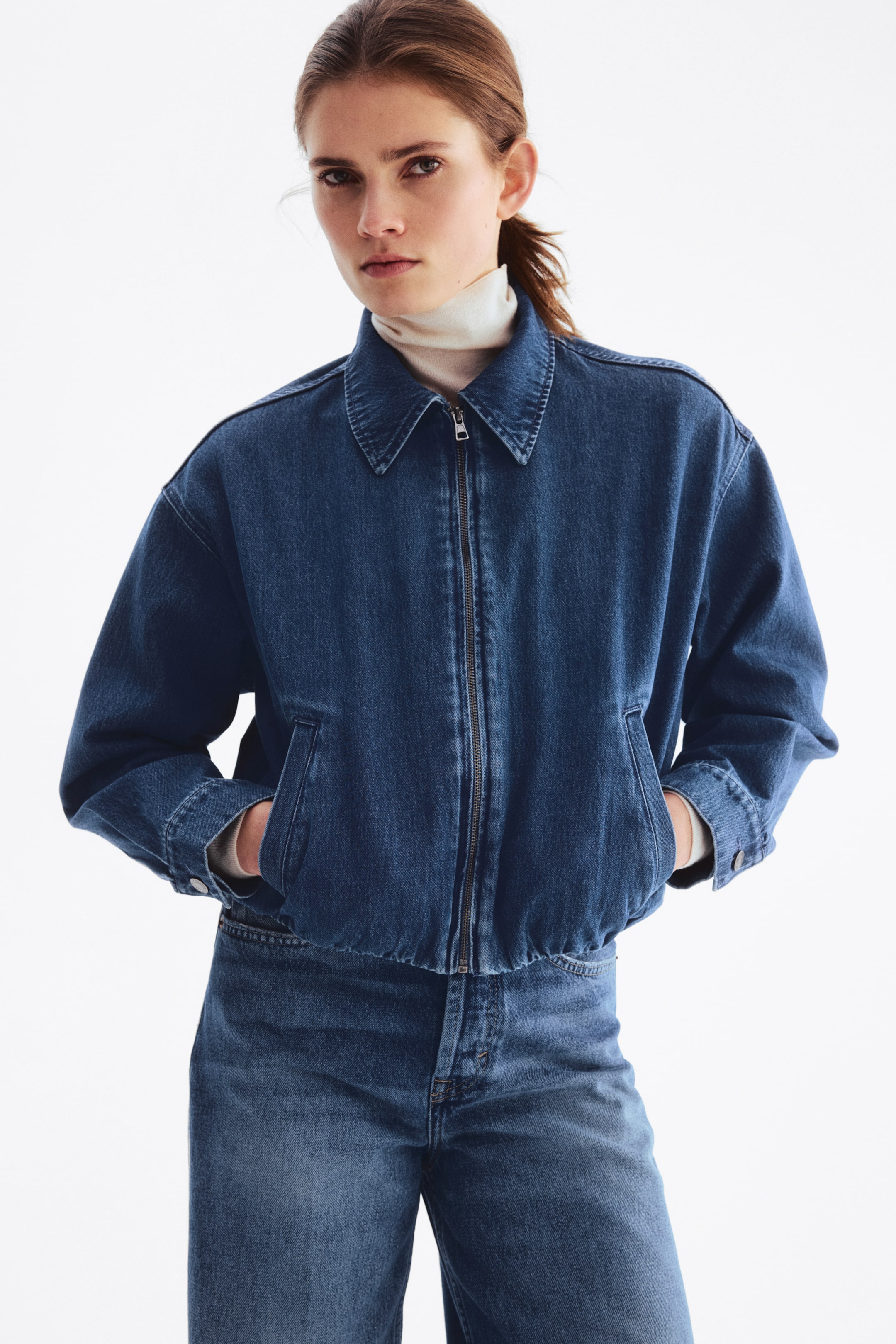 View larger image: The model is wearing a short, oversized denim jacket with a collar, zippered front, dropped shoulders, buttoned cuffs, and an elastic hem for a boxy silhouette. Her hands are tucked into the jacket's welt front pockets, and it's layered over a light-colored turtleneck, paired with matching denim jeans.