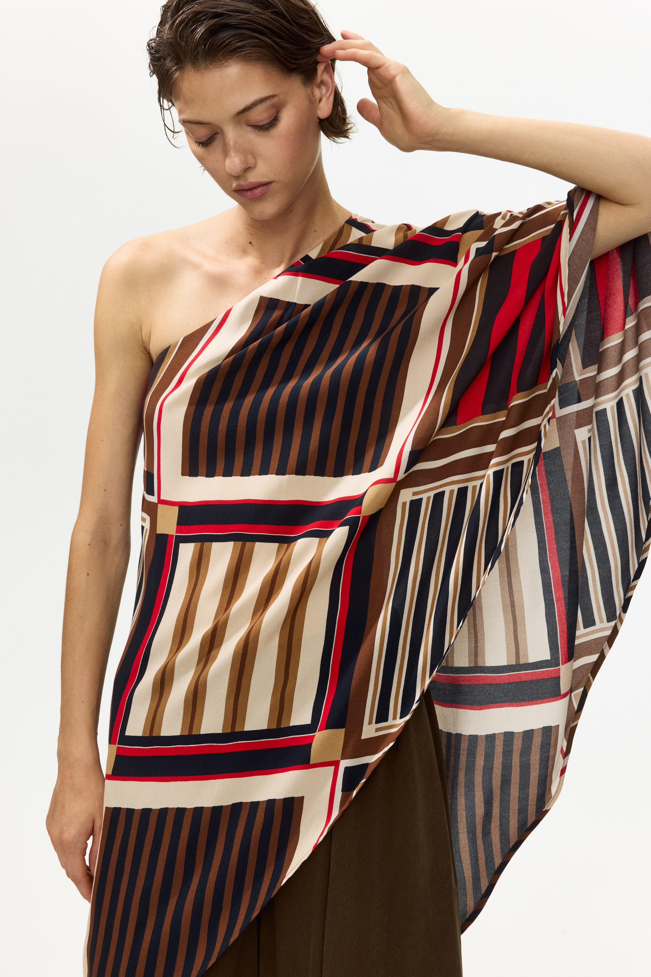 View larger image: A one-shoulder garment showcases a geometric striped pattern in beige, light brown, dark brown, navy, and red, elegantly draping over the torso and one arm, layered atop a dark olive garment.