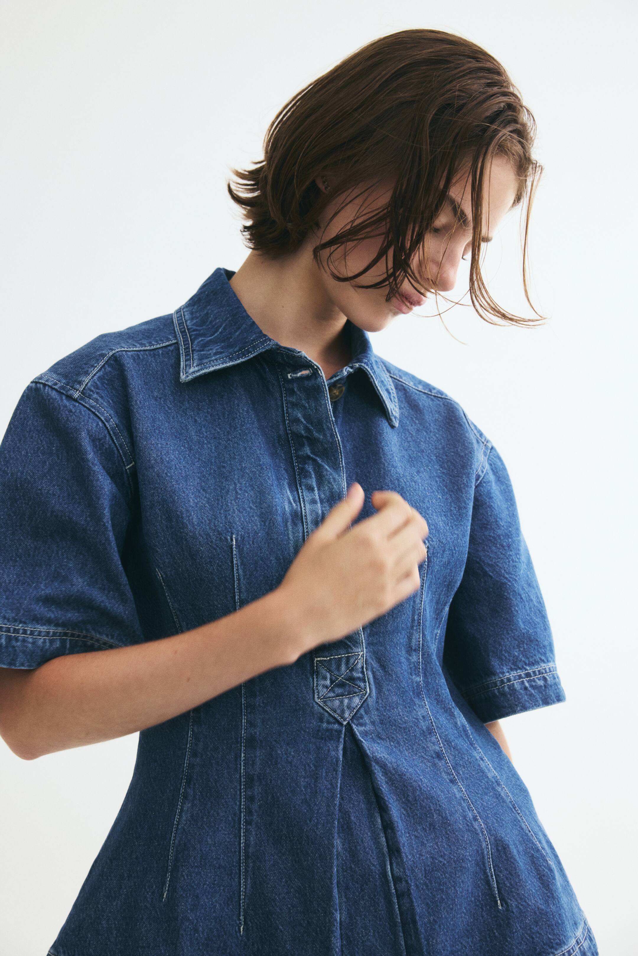 View larger image: The model is wearing a short-sleeved denim shirt. It features a collar with a visible button on the collar stand, a concealed button placket, an inverted box pleat at the front hem, and shaping waist darts at the front. The model is resting one hand on the upper front of the shirt.
