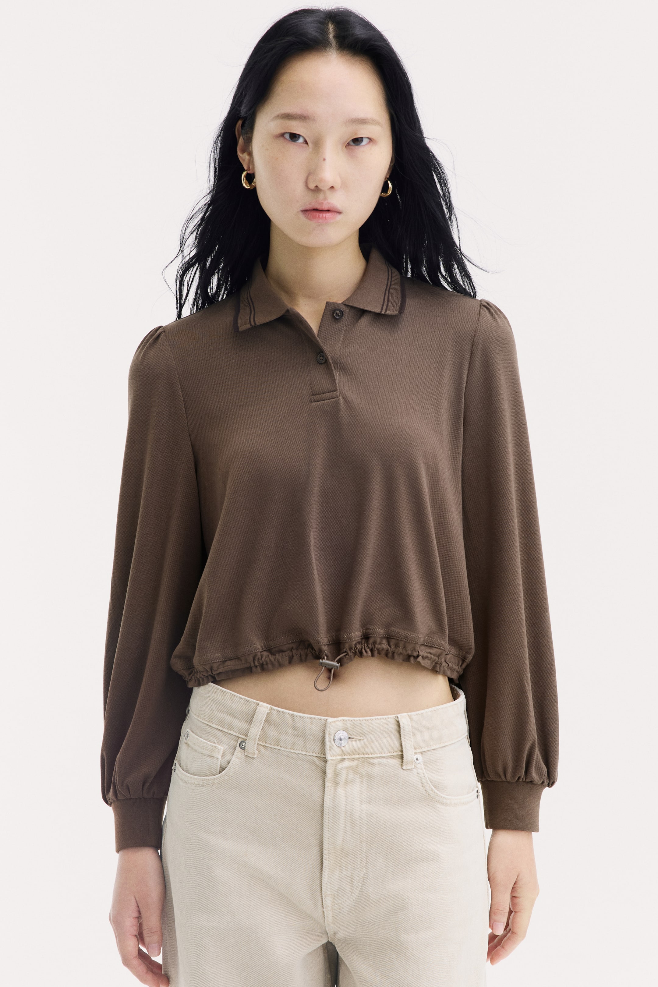 View larger image: The model is wearing a brown polo shirt featuring a ribbed collar, button placket, long puff sleeves with ribbed cuffs, and a cropped drawstring hem with a cord stopper. The shirt is styled with light beige denim jeans.