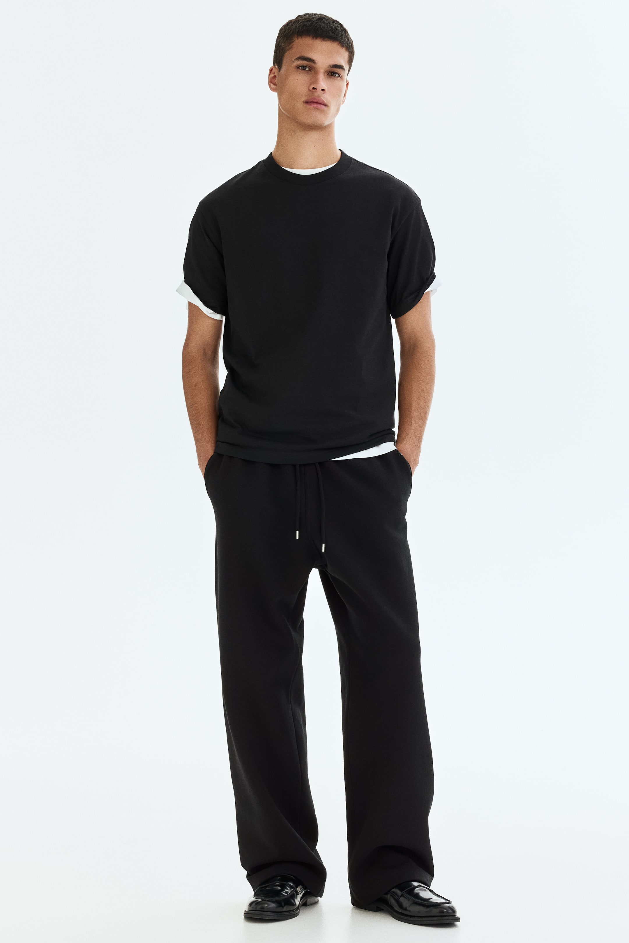 Grösseres Bild ansehen: Schwarze Jogginghosen aus einem schweren Sweatshirt-Stoff werden vom Model getragen. Sie zeichnen sich durch eine lockere Passform, einen Kordelzug an der Taille und Seitentaschen aus, in denen die Hände des Models platziert sind.