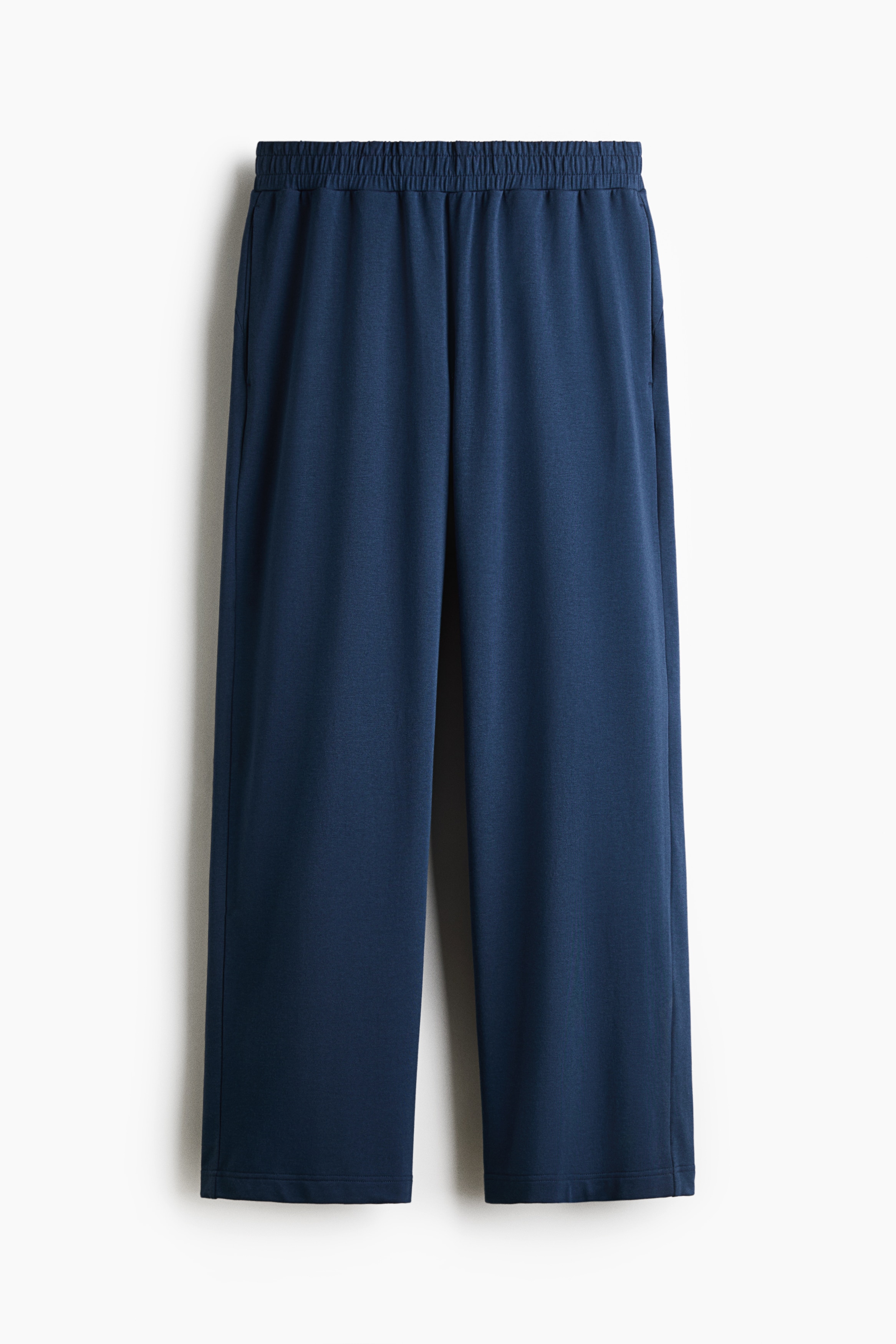 Un pantalon de sport bleu à coupe ample et jambes larges est porté par le modèle, qui se tient de profil avec une main glissée dans une poche latérale. Le pantalon est agencé avec un haut bleu à manches longues assorti et des sneakers foncées.
