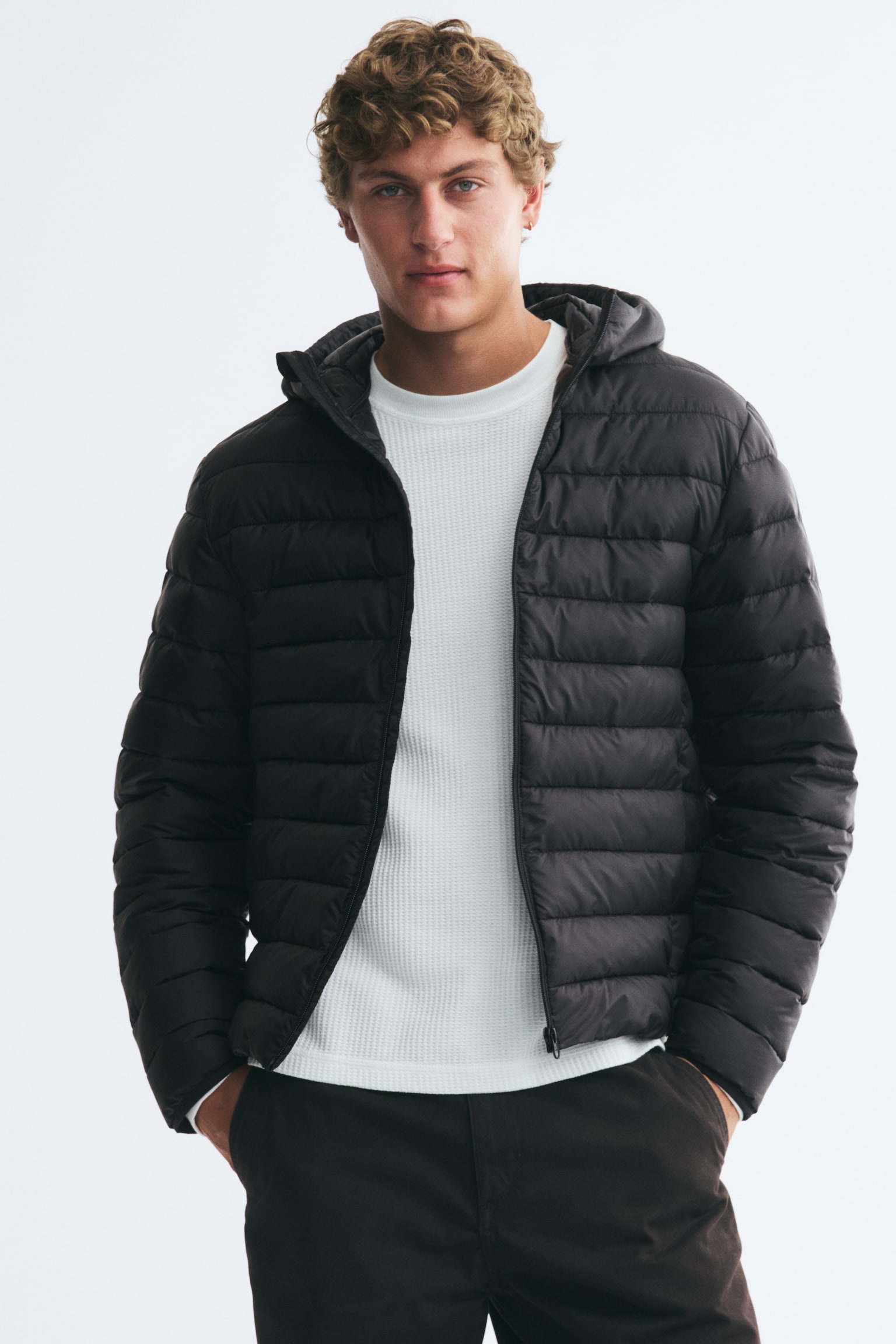 A black front-facing puffer jacket is featuring horizontal quilting throughout its body and sleeves. It is designed with an attached hood, a full-length central zipper closure, and elasticated cuffs. - 1