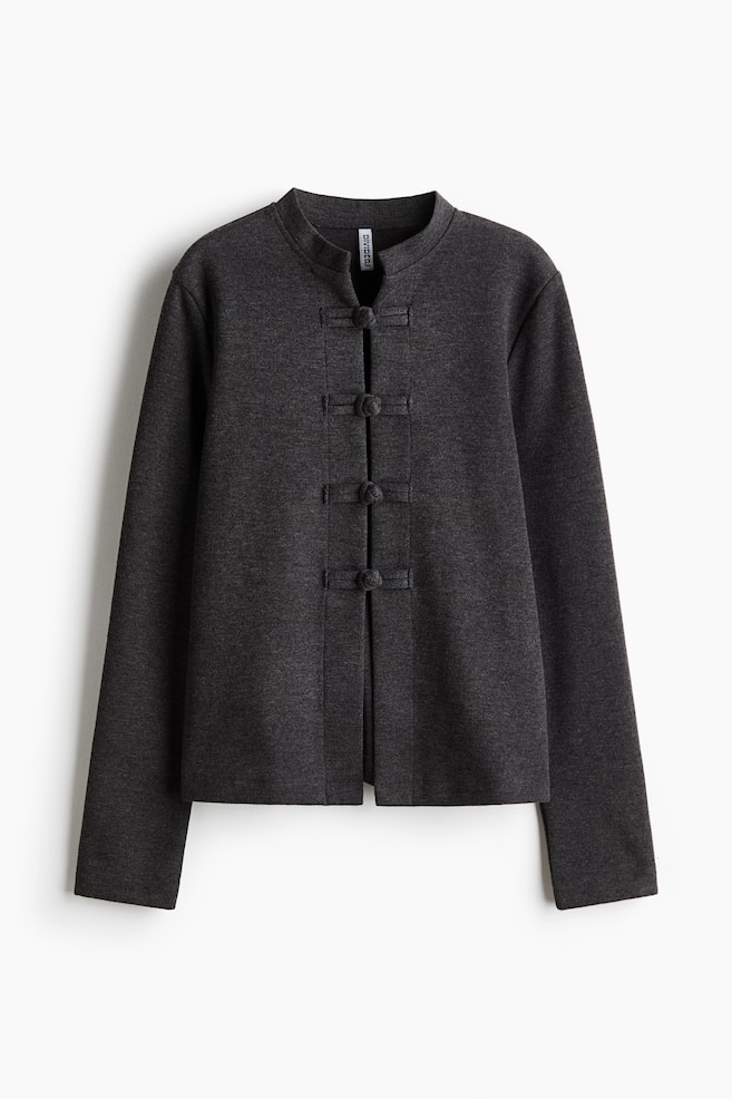 Eine dunkelgraue Strickjacke wird in einer frontalen Ansicht mit einem niedrigen Stehkragen, langen Ãrmeln und einem geraden Saum gezeigt. Vier passende KnotenschlÃ¼sse aus Stoff sichern die vordere Ãffnung des fein gestrickten KleidungsstÃ¼cks. - 1
