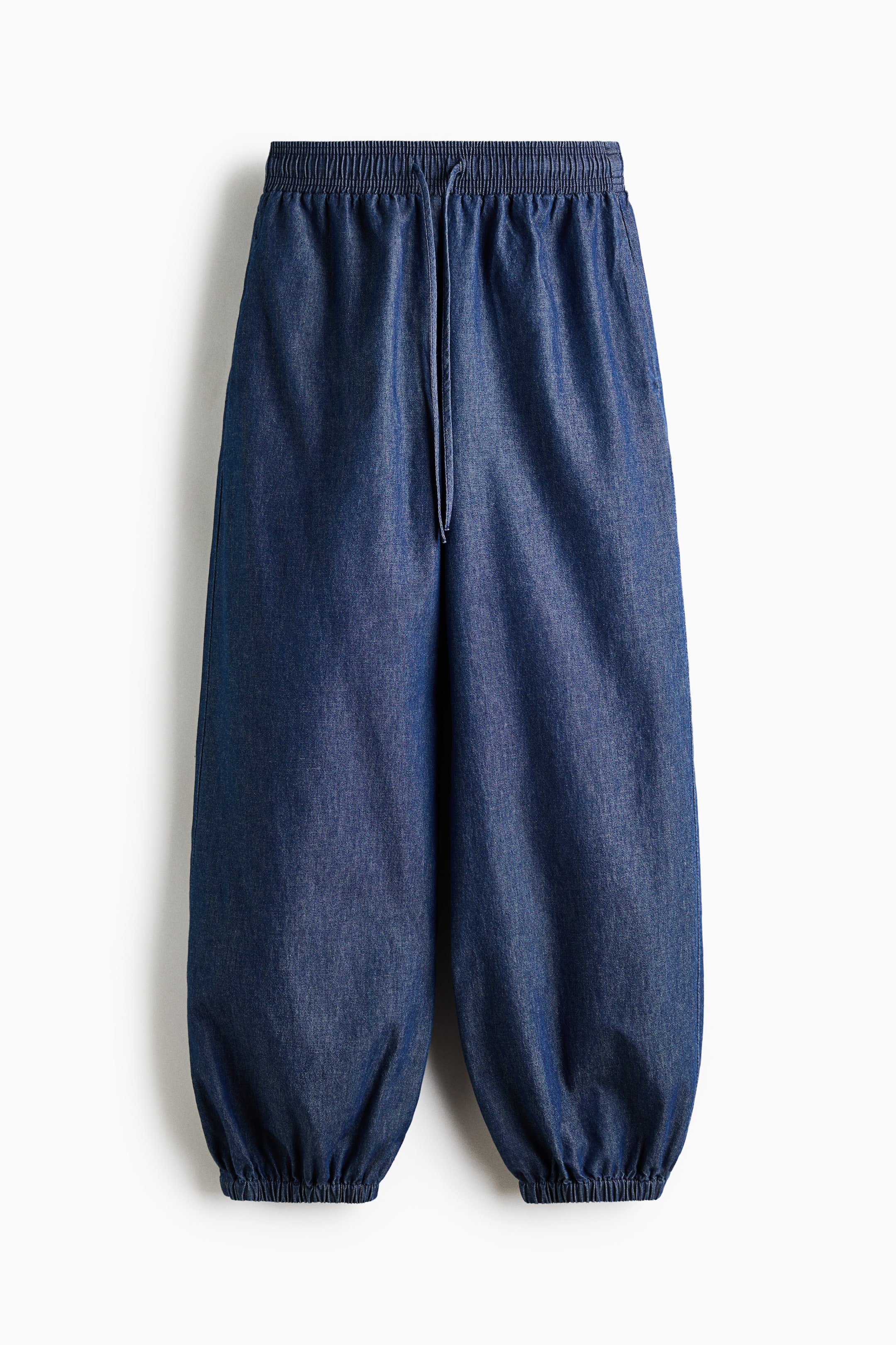 Xem ảnh lớn hơn: Quần denim màu xanh đậm có cạp thun với dây buộc trung tâm phía trước. Ống quần rộng, phồng được hoàn thiện với cổ tay thun, tạo dáng tụ lại ở mắt cá chân.