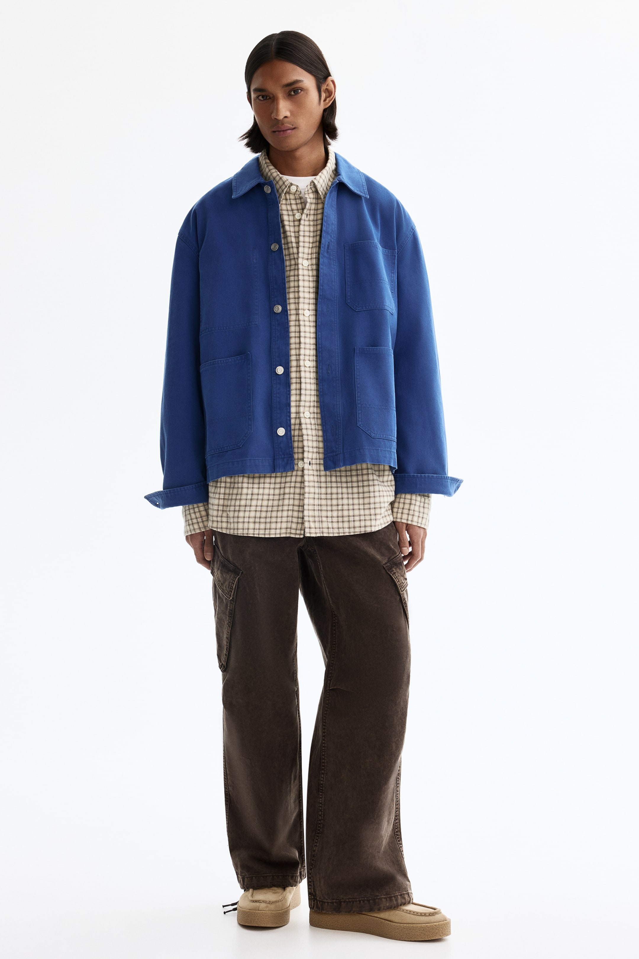 View larger image: A blue workwear jacket with a collar, button-front and four patch pockets is worn open with cuffed sleeves over a light plaid button-up shirt. Dark brown cargo pants, featuring side cargo pockets, are paired with beige moccasin-style shoes, showcasing the ensemble from the front.