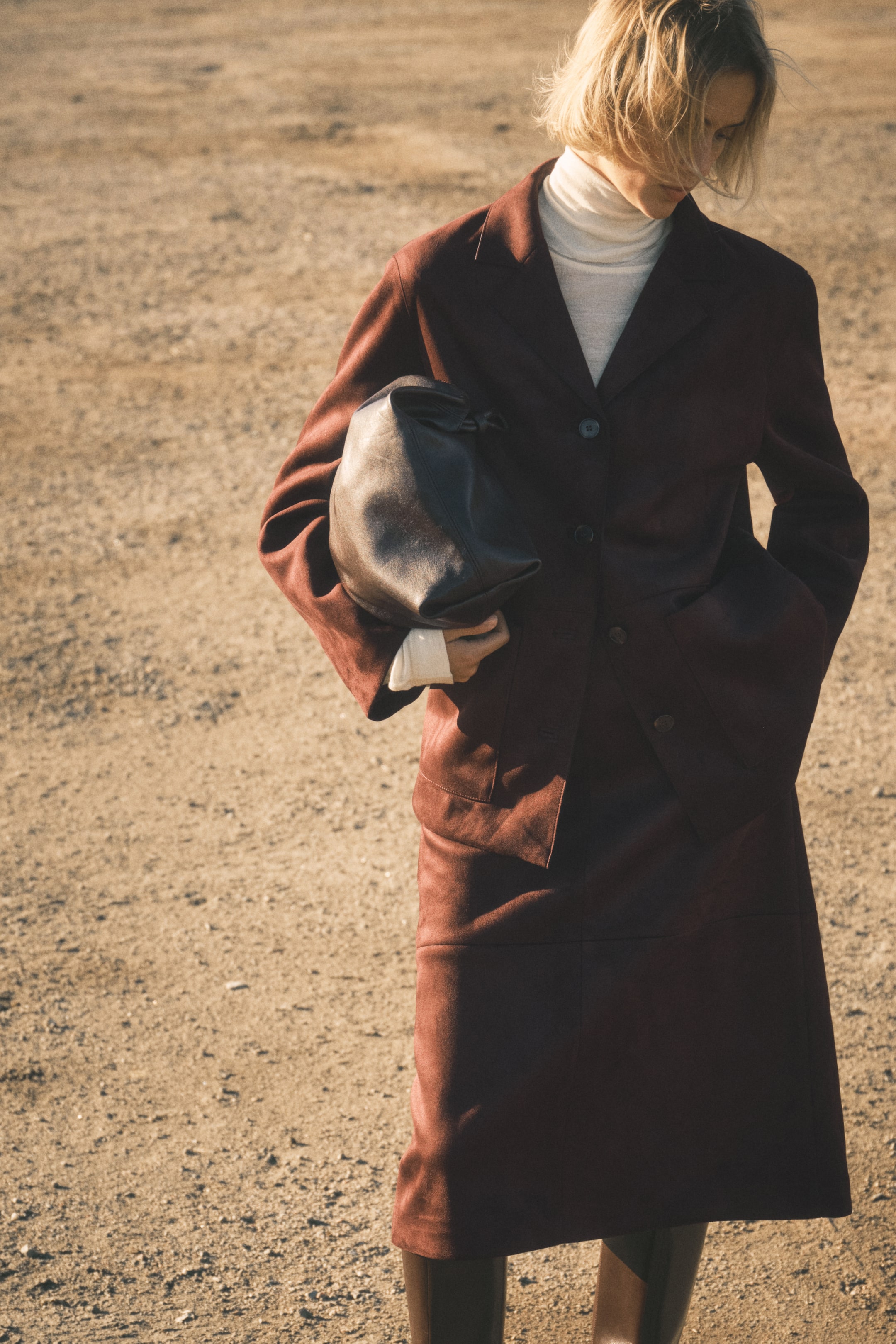 Vezi imaginea mai mare: Un costum din imitație de piele întoarsă bordo, prezentat dintr-un unghi din față, prezintă o jachetă cu un singur rând de nasturi cu reveruri în crestătură și o fustă dreaptă asortată. Un pulover cu guler rulat crem este purtat sub jachetă, și un plic din piele moale și închisă la culoare este ținut în mână, cu cizme lungi și întunecate vizibile sub fustă.