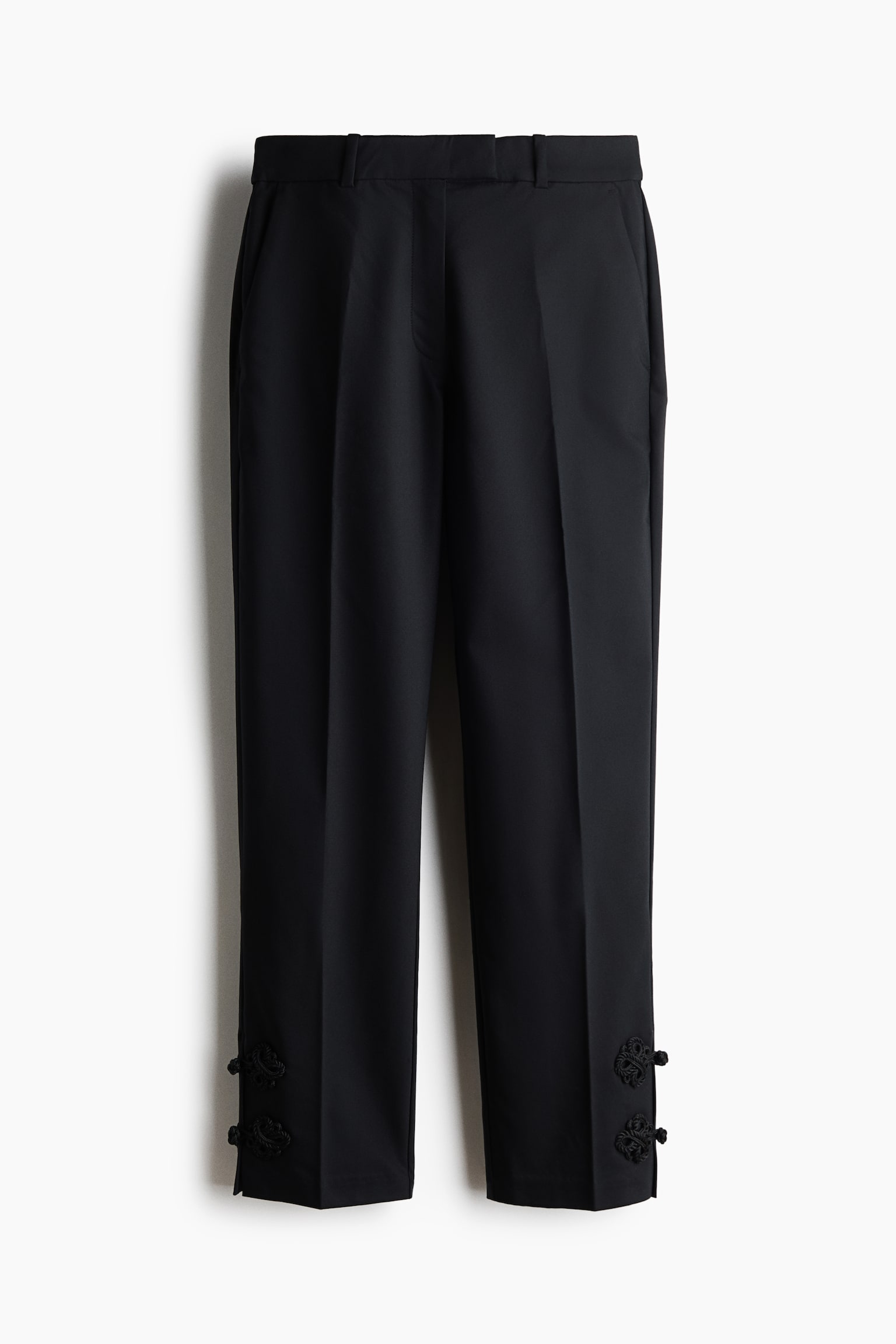 A pair of black tailored trousers is shown from the front, featuring a structured waistband with belt loops and sharp creases running down each leg. The cropped hems display two decorative black braided frog fastenings on the outer sides.