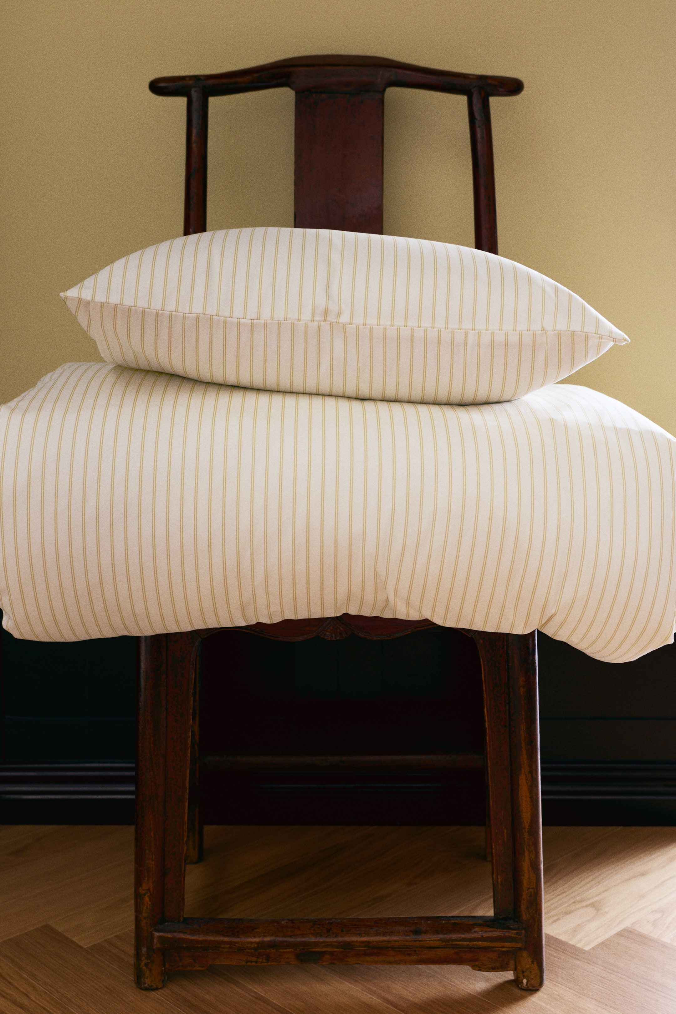 Voir une image plus grande: Un ensemble de housse de couette en satin de coton crème présentant un fin motif à rayures beiges verticales est montré de face, avec la housse de couette pliée drapée sur une chaise en bois foncé et la taie d’oreiller assortie posée dessus.