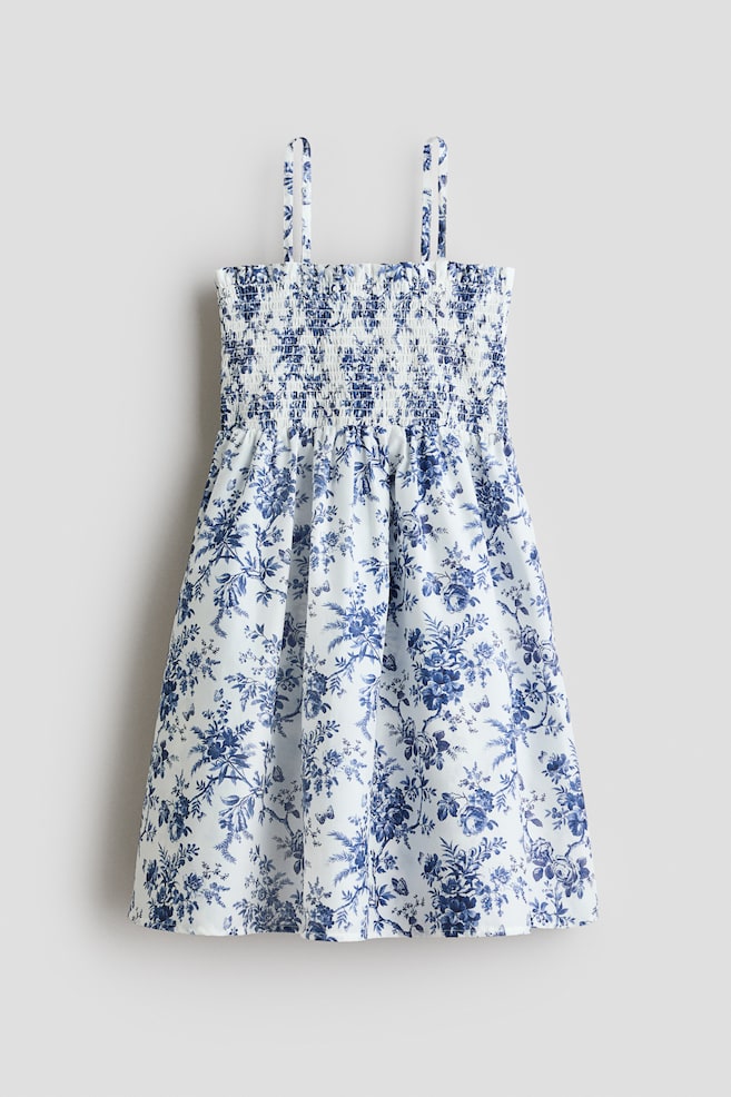 El vestido presenta un patrón floral azul sobre un fondo blanco, visto desde el frente. Cuenta con un corpiño fruncido con tirantes ajustables y estrechos, una costura recogida en la cintura y una falda suavemente acampanada. - 1
