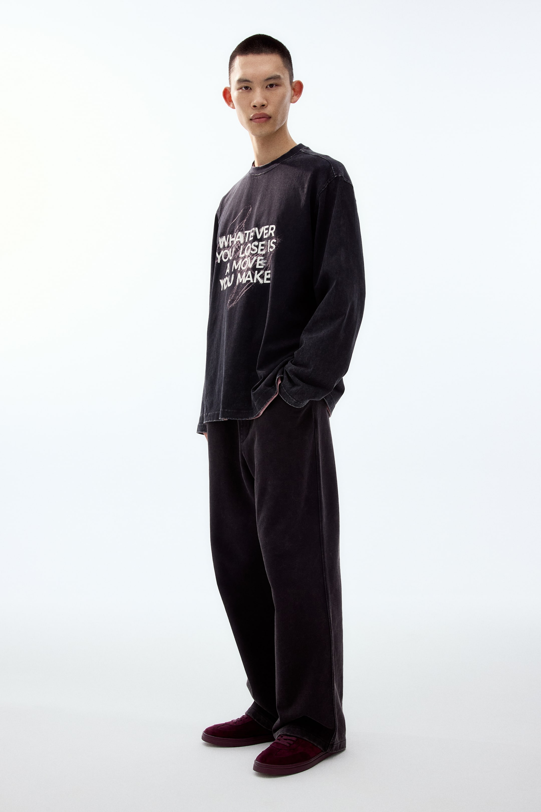 拡大画像を表示: 黒のウォッシュ加工が施された長袖クルーネックシャツ。胸元に白いグラフィックテキスト「WHATEVER YOU LOSE IS A MOVE YOU MAKE」が入っており、やや斜め前方からの視点で表示されています。黒のワイドパンツとダークレッドのロートップスニーカーでコーディネートされています。