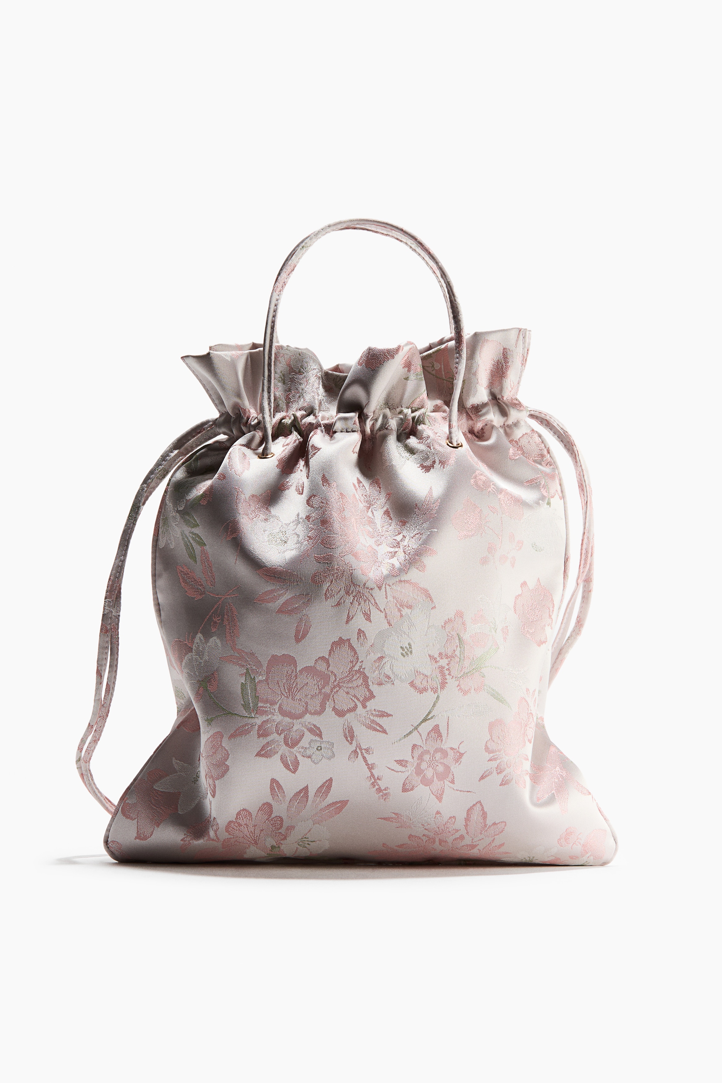 Un petit sac seau en satin doux Ã  motif floral rose et blanc est tenu par le mannequin. Le sac prÃ©sente de fines anses supÃ©rieures et une fermeture Ã  cordon froncÃ©e.