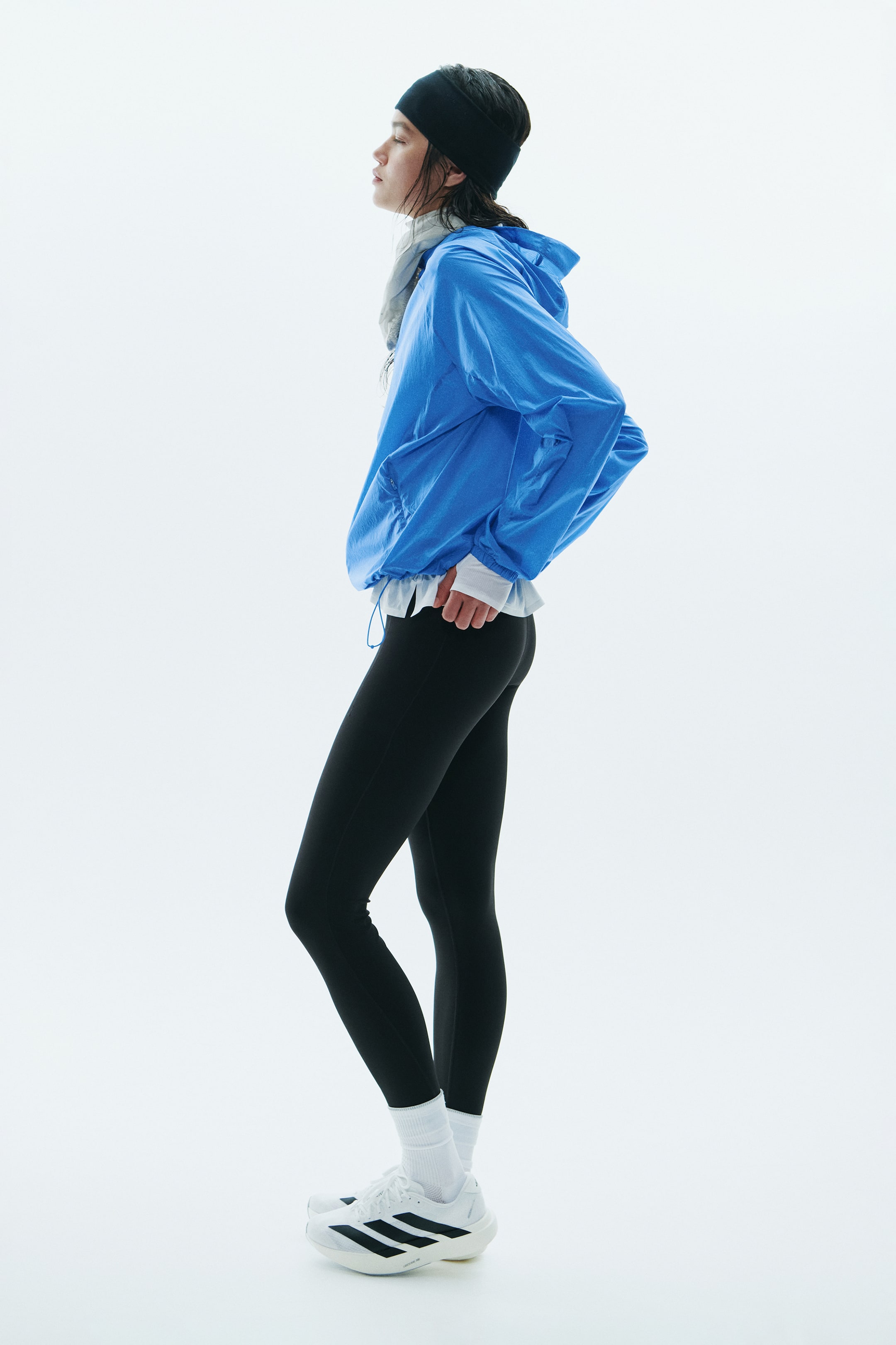 Größeres Bild anzeigen: Eine blaue Laufjacke mit entspannter Passform wird vom Model getragen und über einem weißen Langarmshirt sowie einer schwarzen Leggings kombiniert. Die leichte Jacke verfügt über eine Kapuze, elastische Ärmelbündchen und einen sichtbaren Kordelzug am Saum.