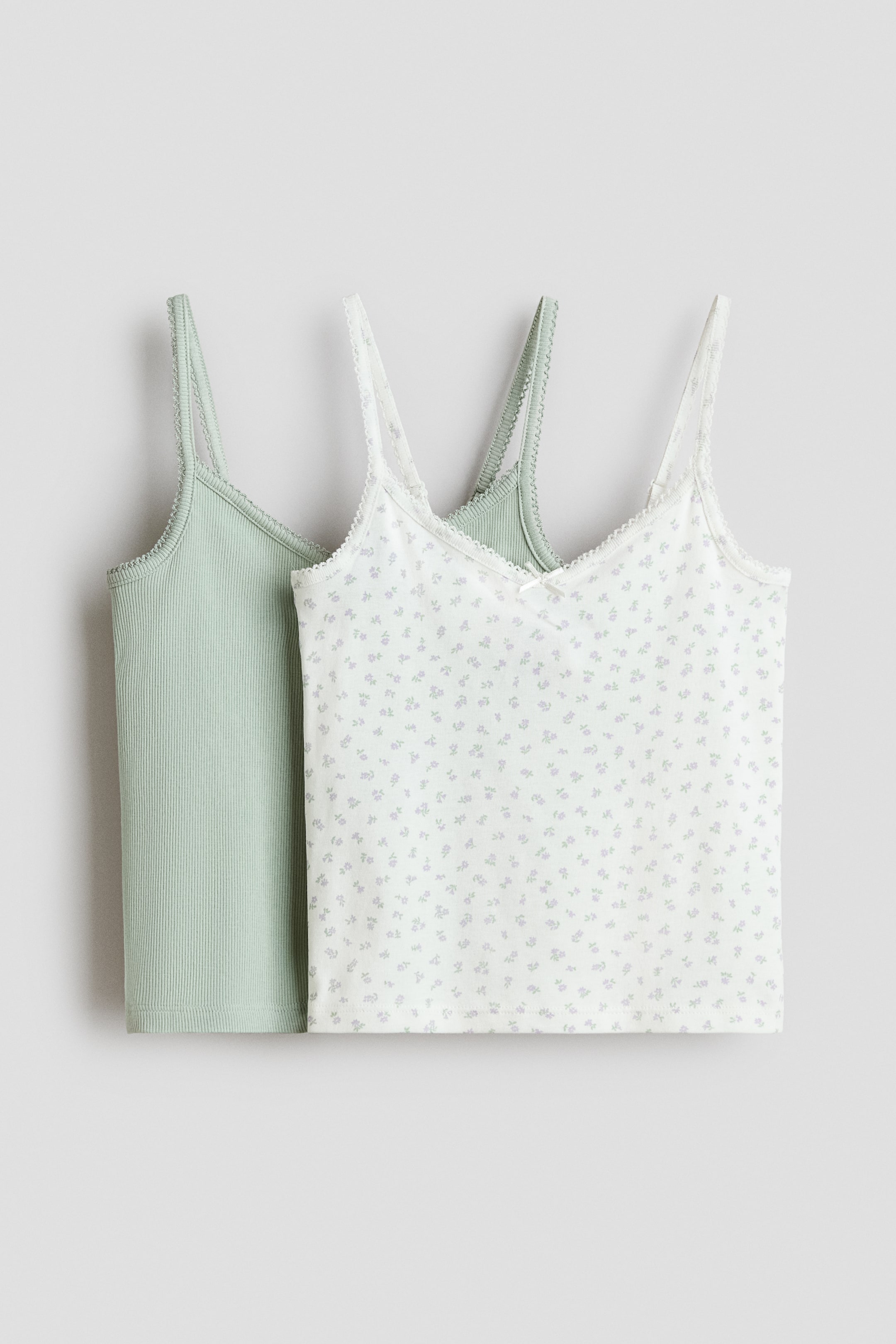 View larger image: Two camisole tops are showcasing their front views. One camisole boasts a light green ribbed knit with scalloped-edge straps and neckline. The other camisole highlights a white fabric adorned with a small blue floral pattern, featuring delicate lace trim along the V-neck and straps, and a small bow at the center front.