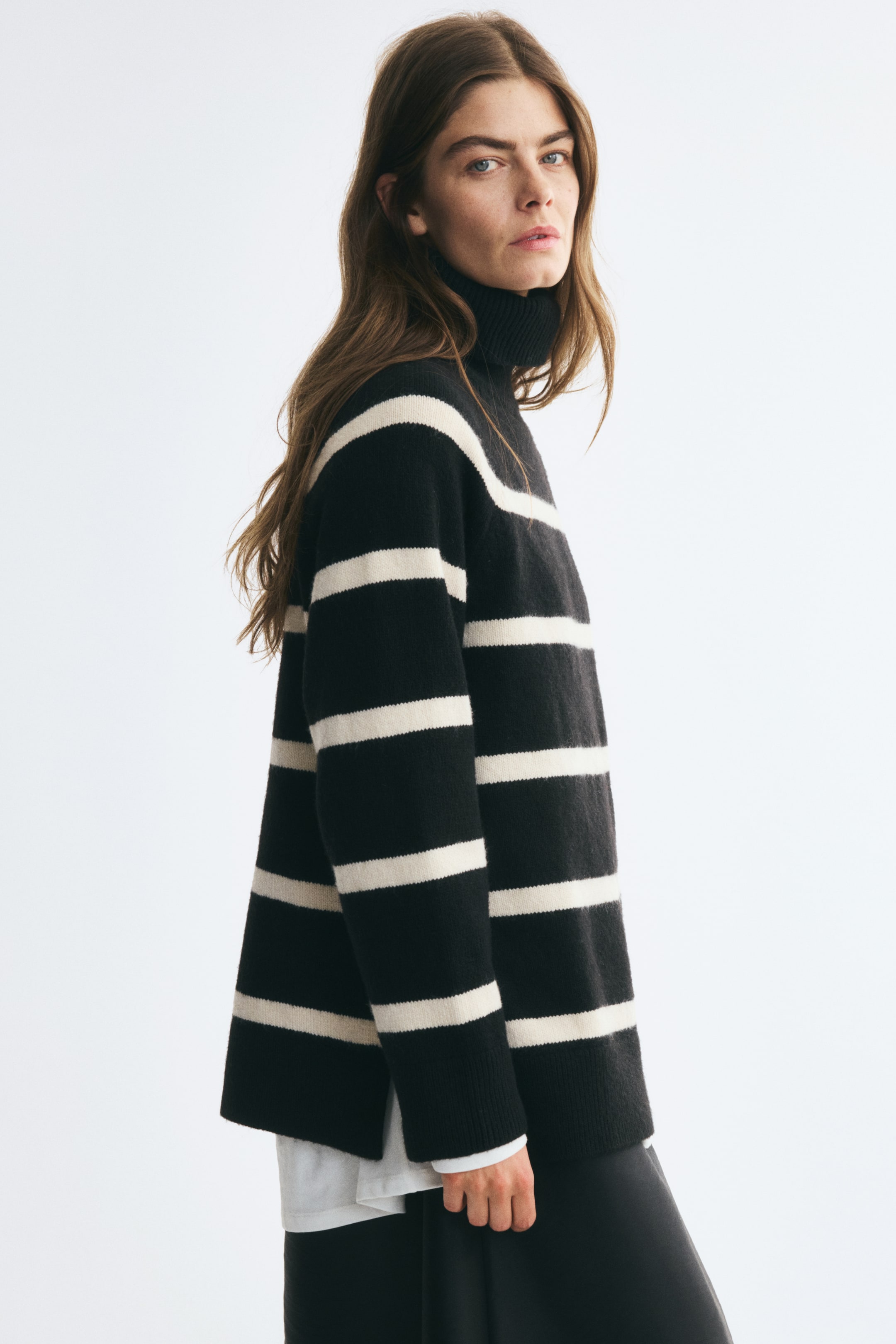 View larger image: An oversized black-and-cream striped knit sweater is worn by the model. The sweater features a ribbed turtleneck, long raglan sleeves, and a ribbed hem with side slits, layered over a white shirt.