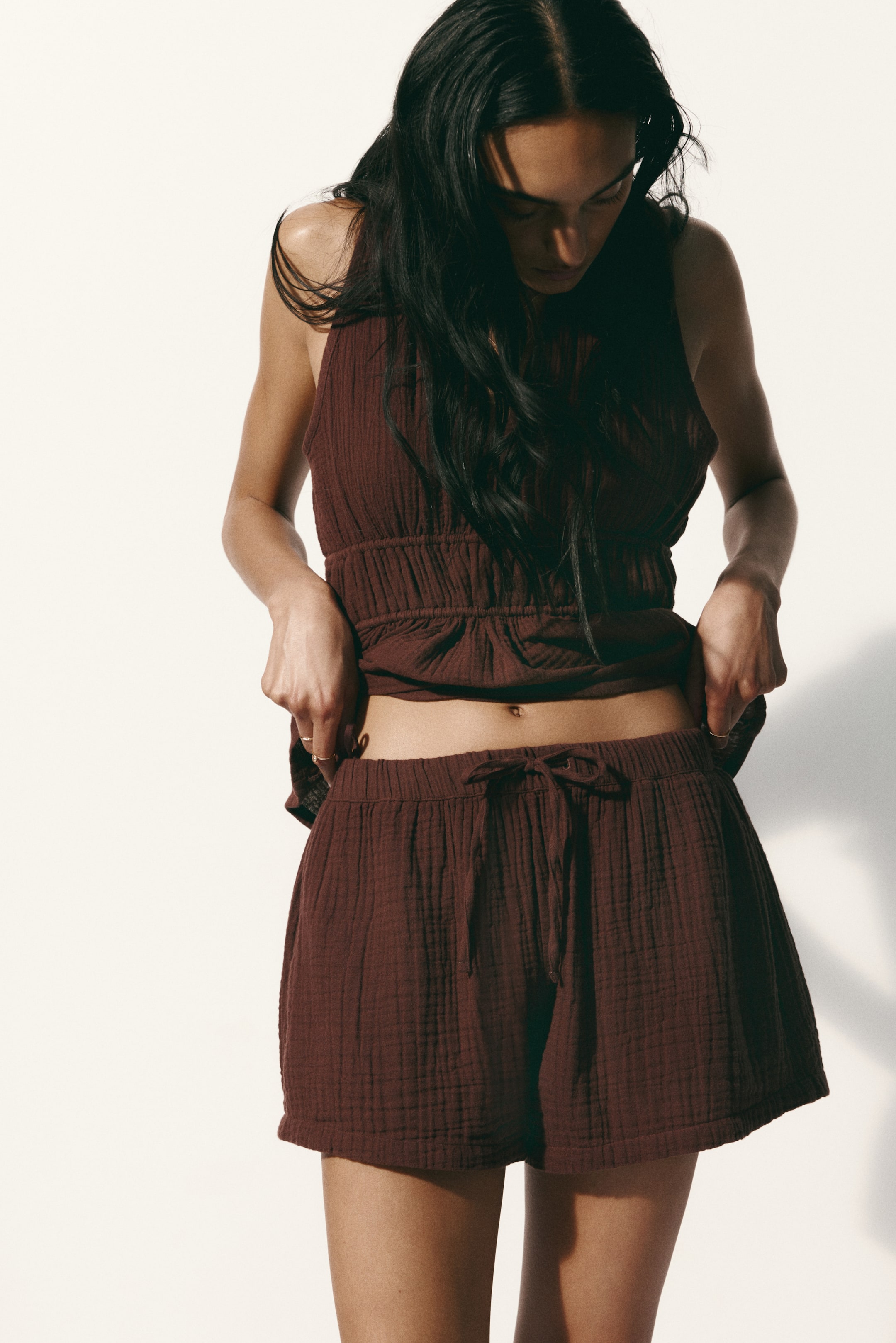 View larger image: A dark reddish-brown crinkled gauze sleeveless top and matching shorts are displayed from a front-facing angle. The top features a gathered, smocked waist, while the shorts are designed with an elastic drawstring waistband. Hands gently pull at the fabric to highlight its texture and fit.