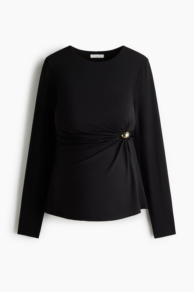 Een zwarte top met lange mouwen wordt van voren getoond, met een ronde hals en asymmetrische plooien aan de zijkant. De gerimpelde stof is vastgezet met een gepolijste, goudkleurige koepelaccent, waardoor zachte gedrapeerde plooien over het voorpand ontstaan. - 1