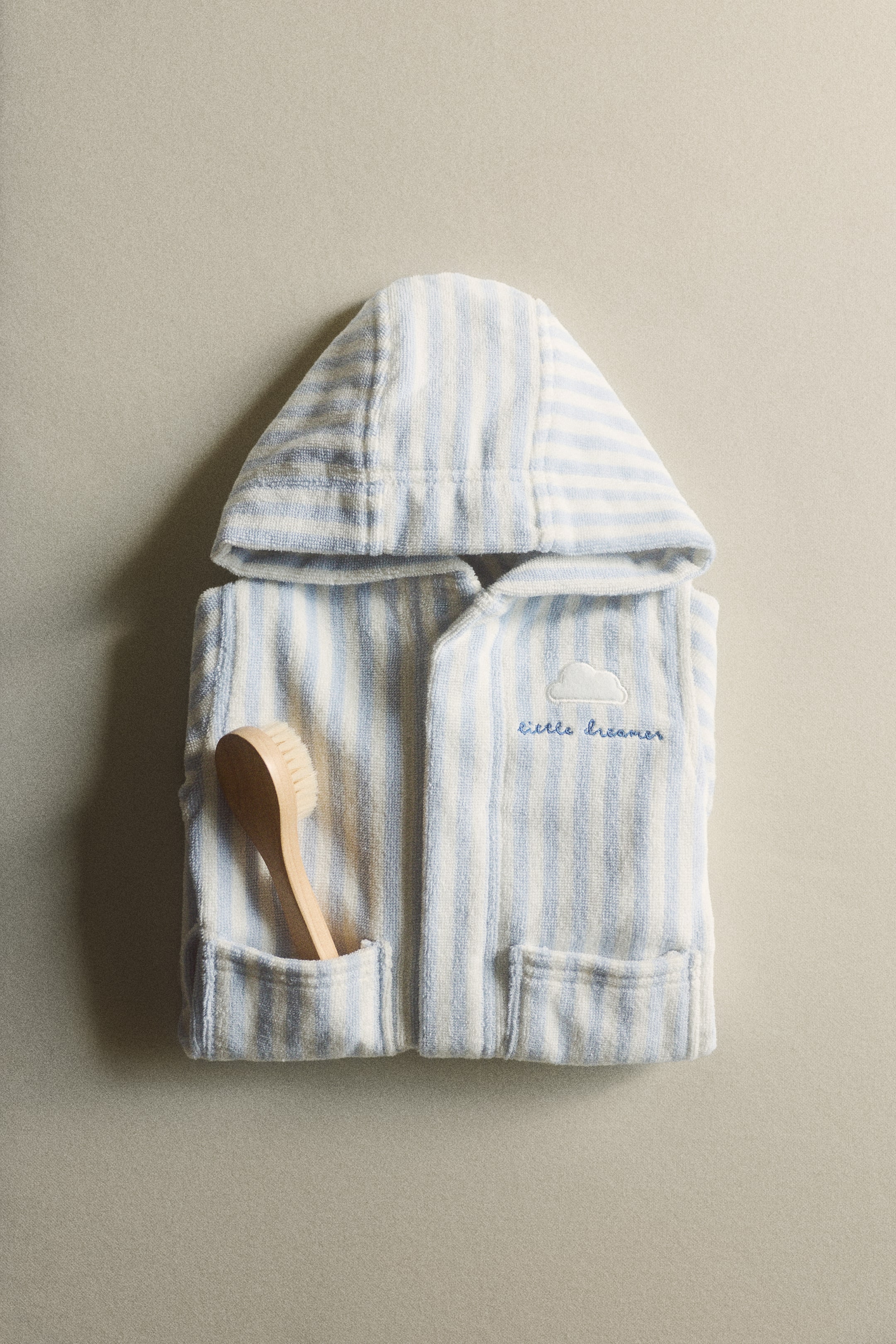 Größeres Bild ansehen: Ein Kapuzen-Babybademantel, gestreift in Hellblau und Weiß, liegt flach und gefaltet. Er verfügt über zwei aufgesetzte Vordertaschen und eine gestickte weiße Wolke mit der Aufschrift "little dreamer" auf der linken Brust, während eine Holzbürste für Babys in der linken Tasche steckt.