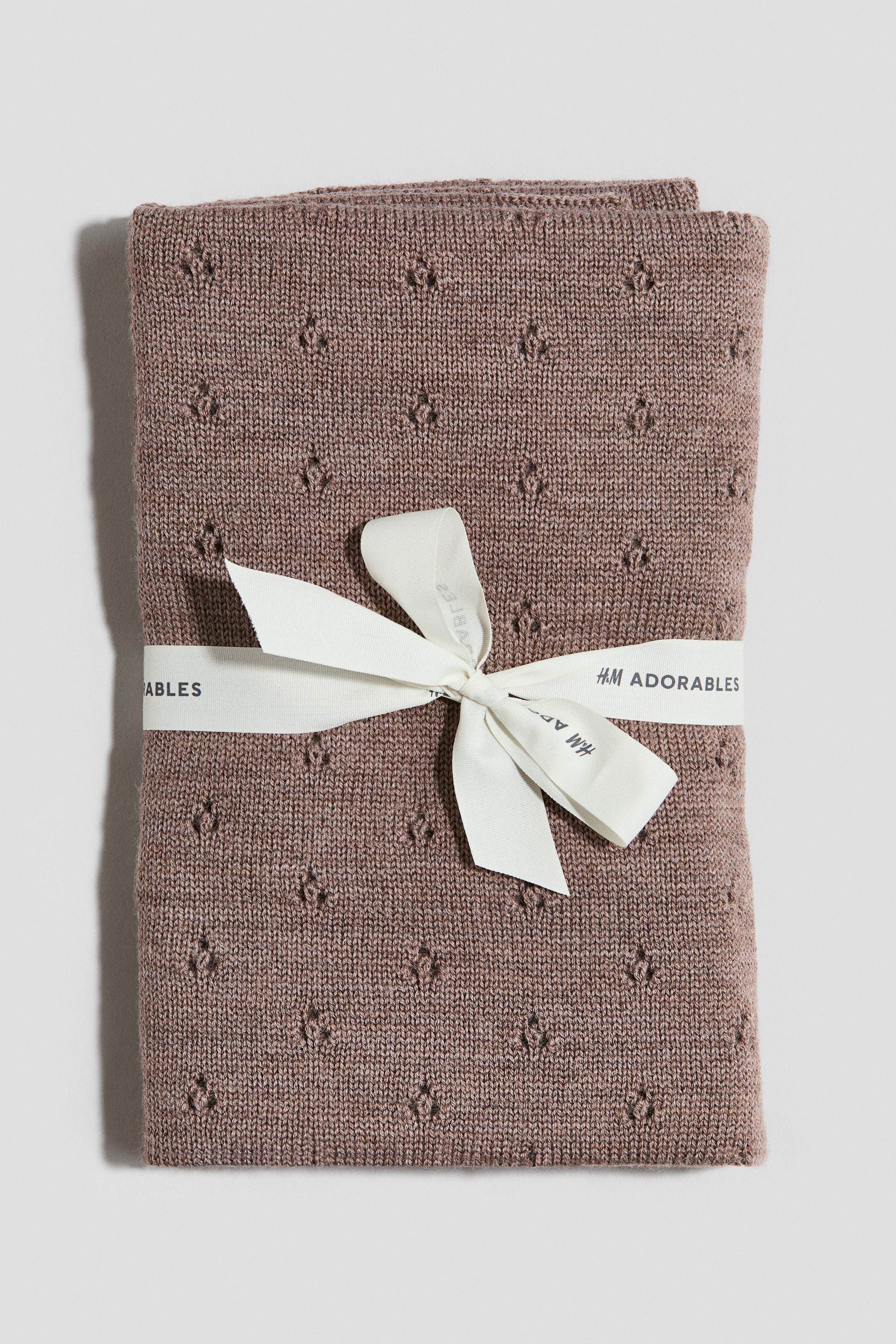 Visa större bild: En dovt rosbrun filt är vikt i en rektangulär form och visar framsidan. Den har ett fint pointelle-stickat mönster över hela ytan, och ett krämfärgat band med varumärket "H&M ADORABLES" är knutet runt mitten i en rosett.