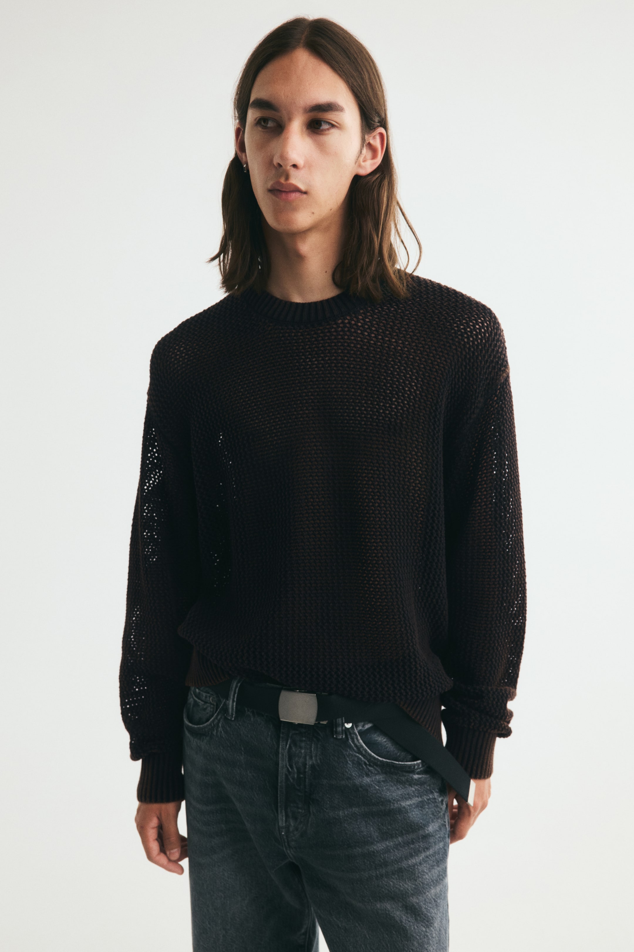 View larger image: A dark brown hole-knit cotton jumper is being worn by the model, featuring a round, rib-trimmed neckline, dropped shoulders, and ribbing at the cuffs and hem. The loose-fitting jumper is partially tucked into dark jeans, revealing a black belt with a silver buckle.