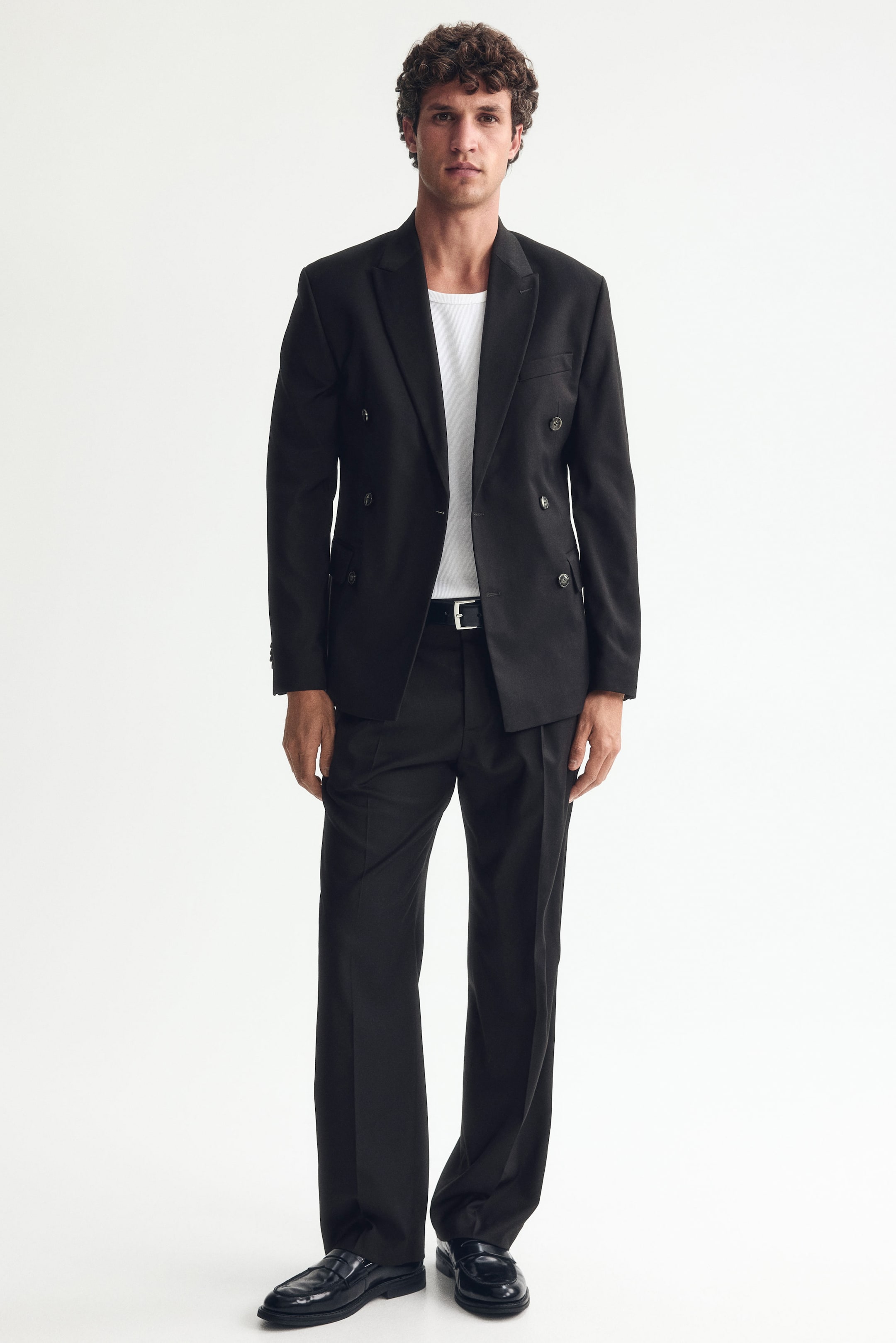 Agrandir l'image: Le mannequin porte un pantalon noir sur mesure avec des jambes droites et des plis. Le pantalon présente des poches latérales inclinées et est maintenu à la taille par une ceinture noire.