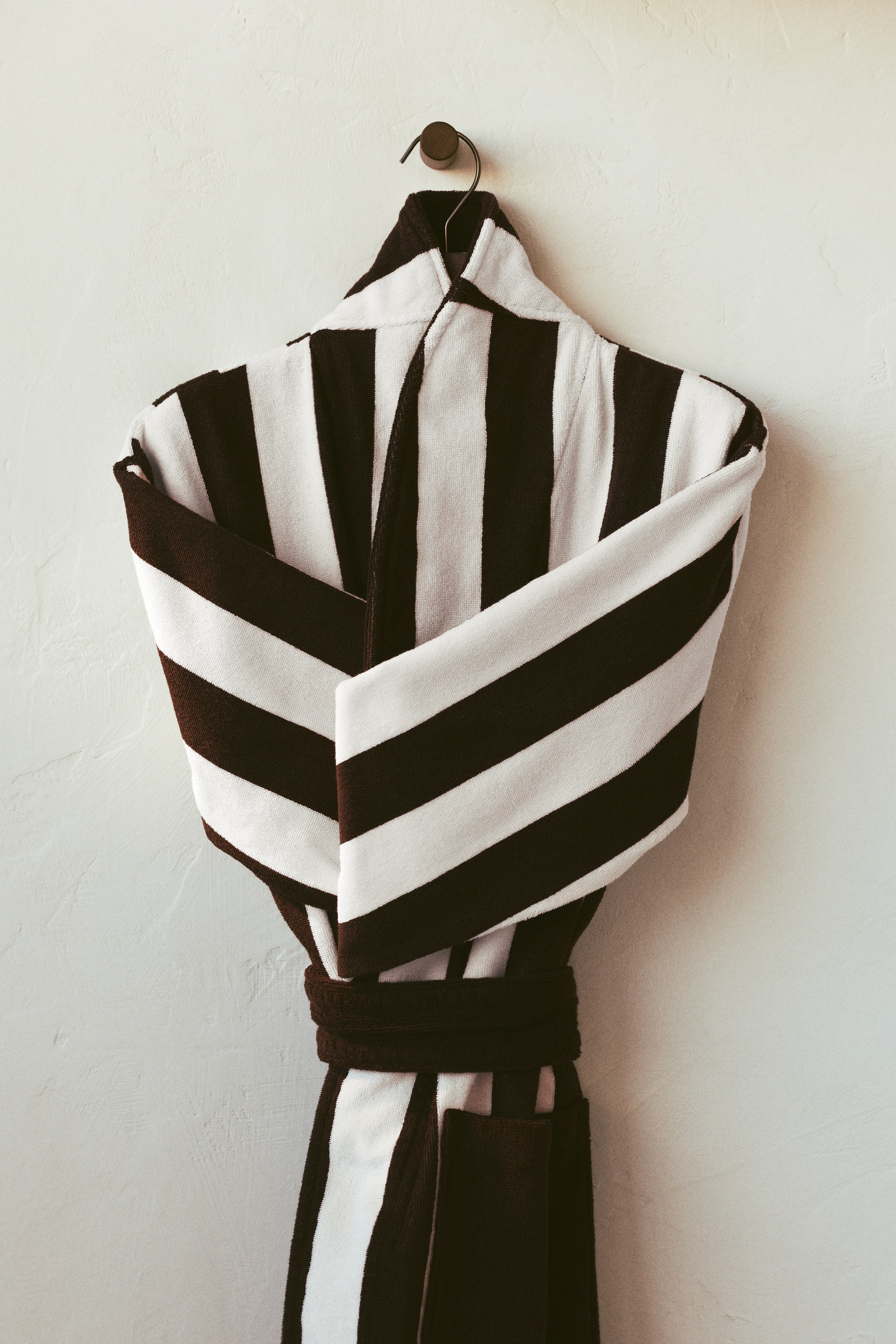 View larger image: A terry cloth dressing gown is displayed, showcasing its front view, with broad vertical stripes in dark brown and white. A dark brown tie belt cinches the waist, and a patch pocket can be seen on the lower right side; its sleeves are neatly folded.