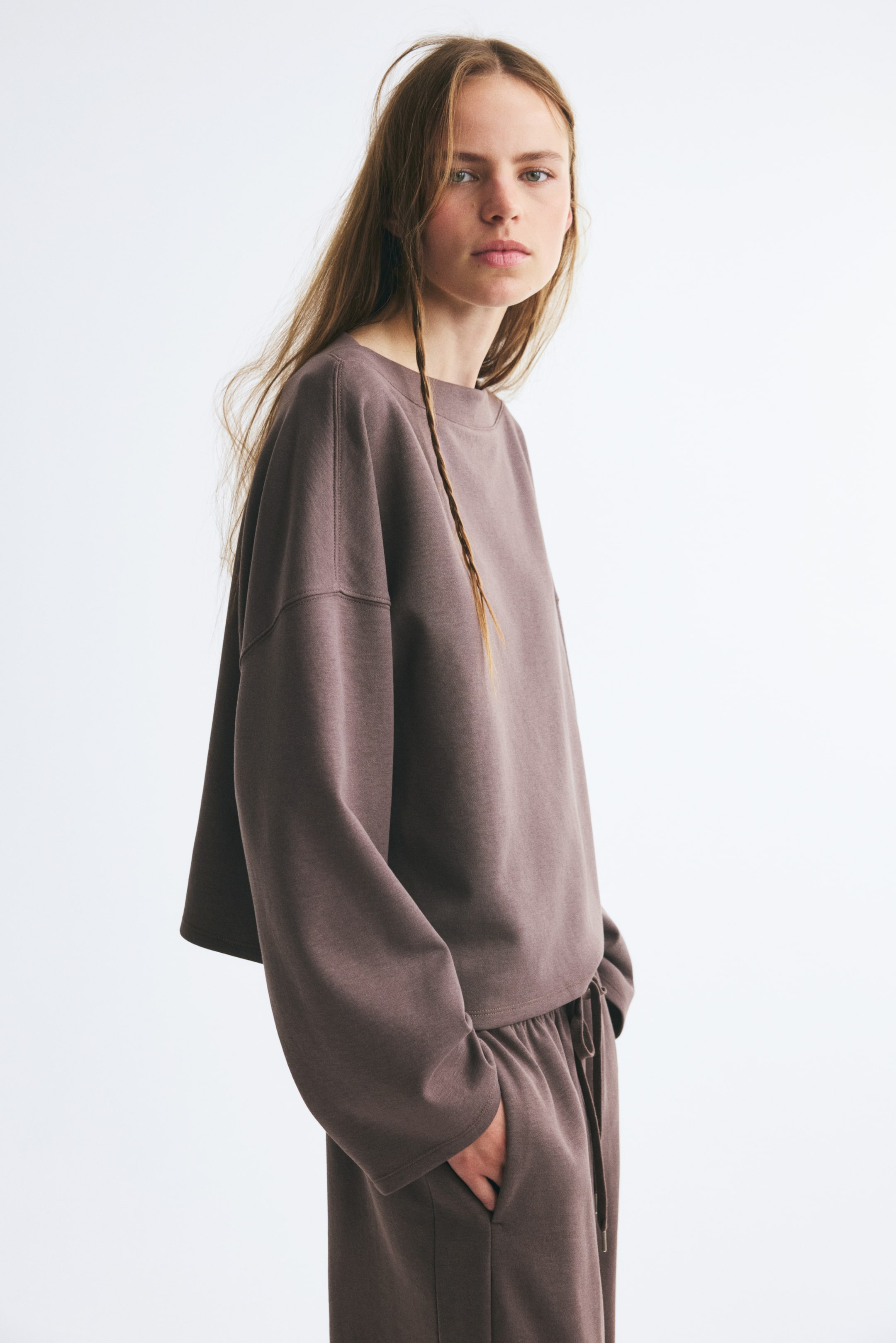 View larger image: A loose-fit, brown fleece sweater featuring a boat neck, low dropped shoulders, and long sleeves is being worn by the model. The sweater is paired with matching drawstring trousers, with the model standing in a side profile view.