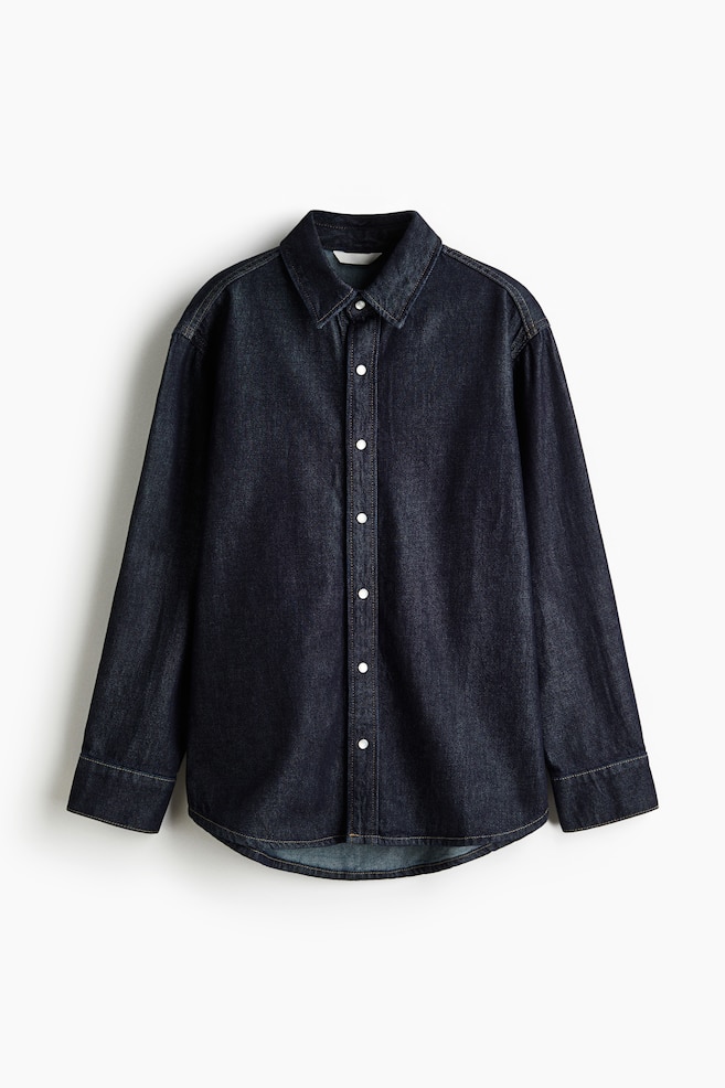 A dark blue denim long shirt is presented from the FRONT. It is featuring a collar, a full-length front placket with white press-studs, and dropped shoulder seams. Long sleeves are ending in press-stud cuffs, and the hem is curved. - 1